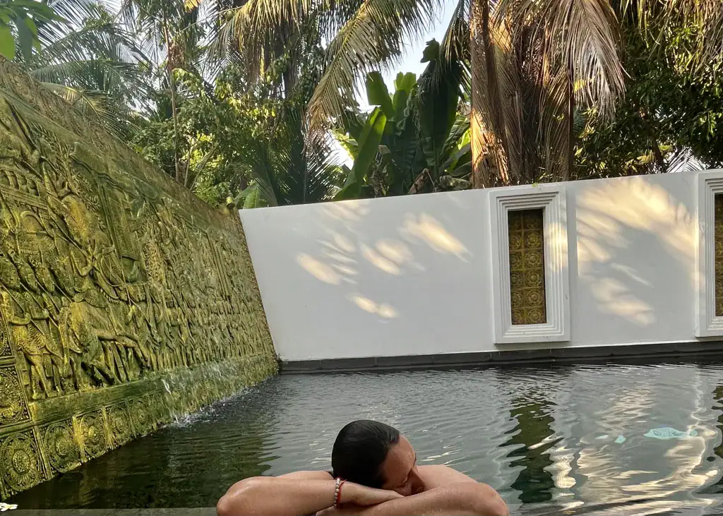 A woman in a swimsuit with wet hair is laying on her crossed arms on the ledge of the pool at The Urban hotel in Siem Reap, Cambodia. One side of the pool is engraved gold figures and the other wall is white with dappled light from the palm trees above the pool shining down.