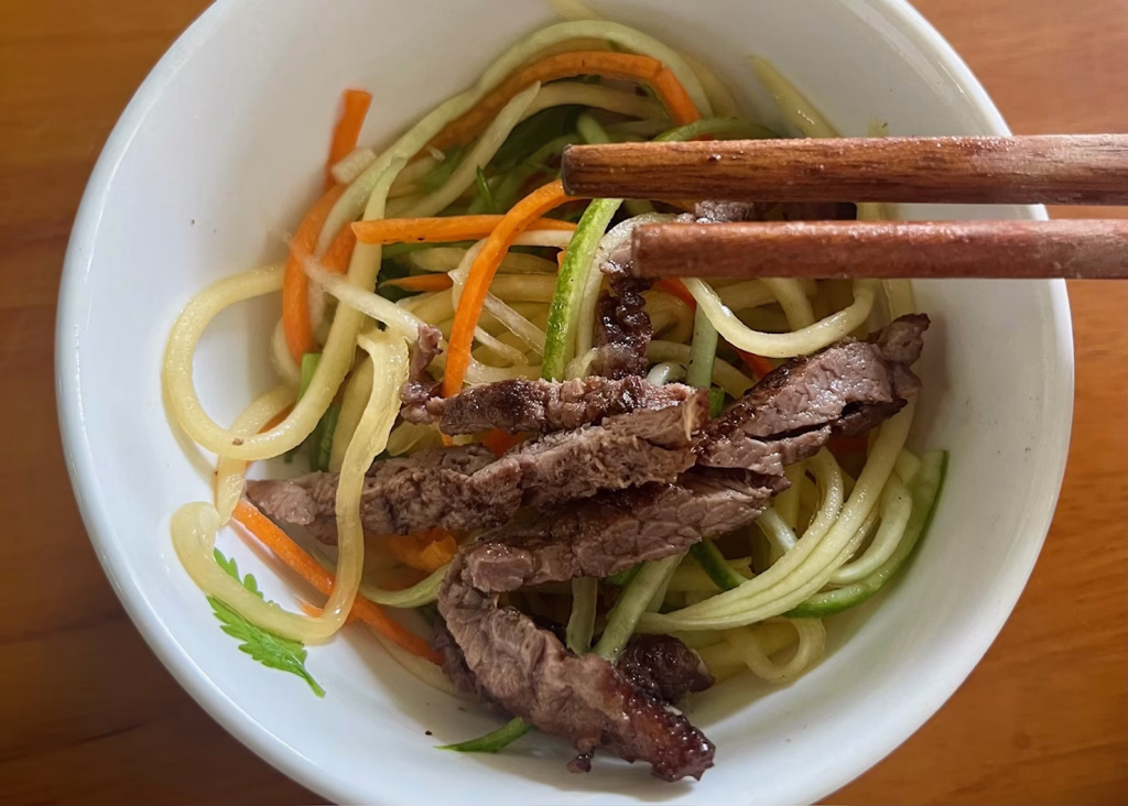 Vietnam 5 A green papaya salad mixed with carrots, herbs, and topped with beef and peanuts is sitting in a white bowl with chopsticks on top.