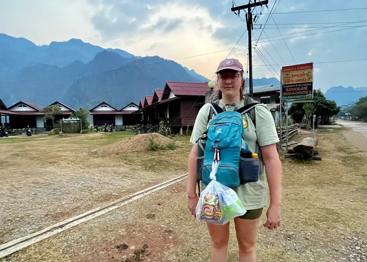 A girl wearing yellow crocs stood outside a bungalow in Laos. She has a backpack on her front whilst she waits for a bus