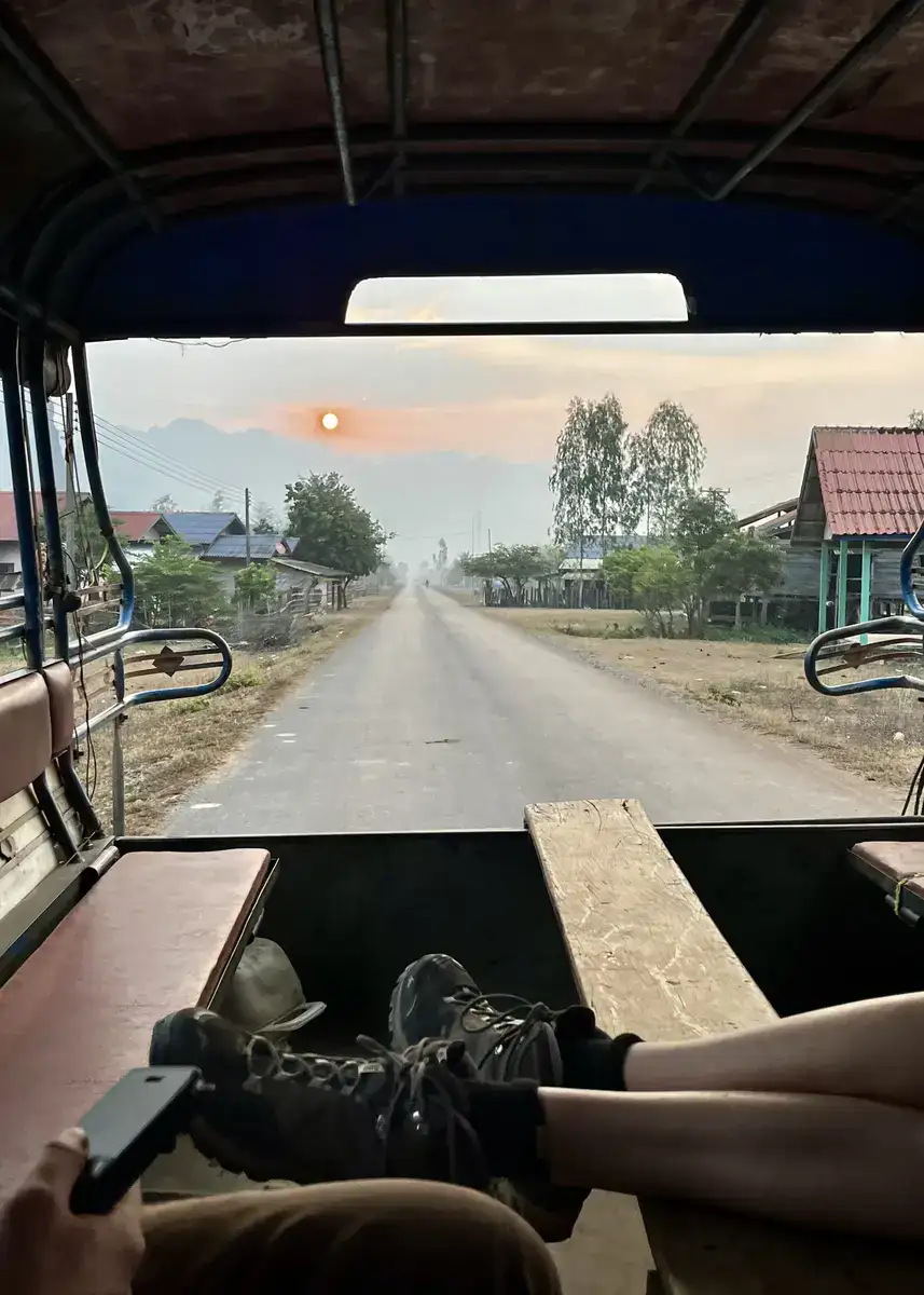 A picture taken from the back of a tuk tuk on the way to Kong Lor Cave by bus in Laos. There is a girl's legs crossed on the benches, as she faces out towards the sunrising over the road