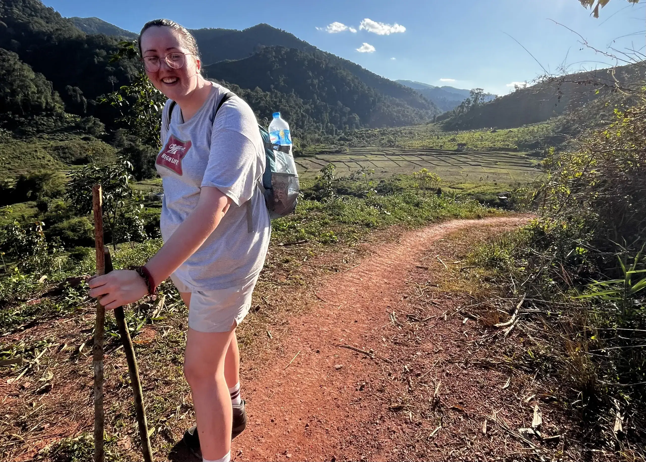 Trekking in Luang Namtha: What to Expect and How to Book 3 A woman wearing a grey tshirt, tan Nike shorts and a backpack is holding two hiking sticks and smiling at the camera. She is standing on a red dirt trail surrounded by green mountains in Nam Ha National Protected Area in Luang Namtha, Laos.