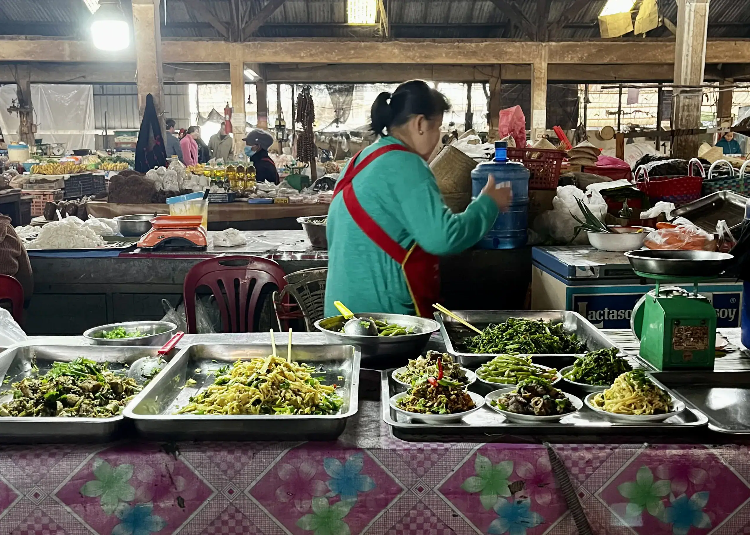 Trekking in Luang Namtha: What to Expect and How to Book 4 A vendor at a morning wet market on the outskirts of Luang Namtha, Laos is preparing premade food for a hiking group to take on the trail. The woman is wearing a jade green sweater and a red apron that crosses in the back.