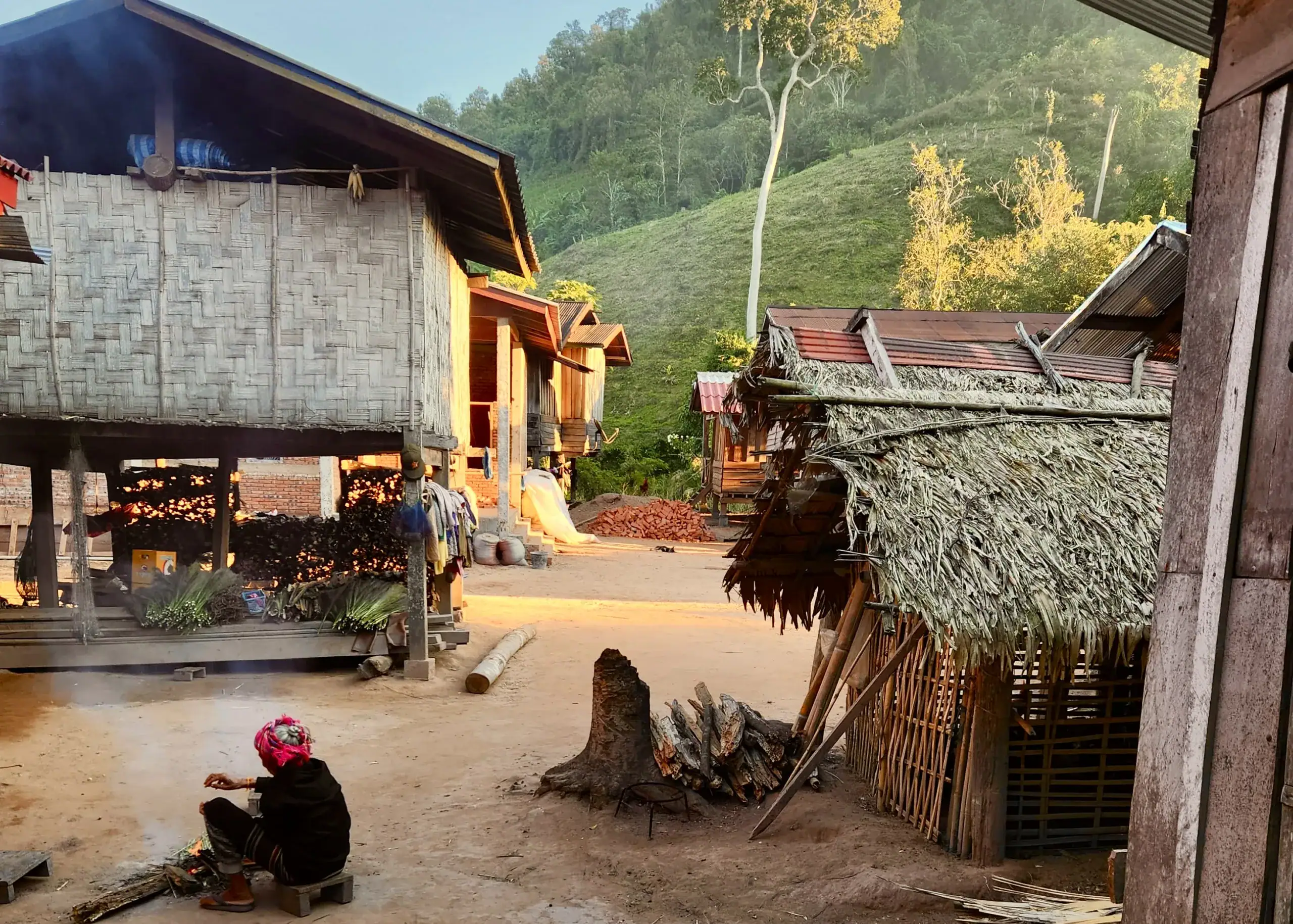 Laos 11 A local village elder is sitting by the fire during a chilly morning in Nam Ha National Protected Area in Luang Namtha, Laos. The houses surrounding the woman are made of bamboo and thatched roofs and she's wearing a bright pink bandana and a black sweater. The green of the mountains surrounding the village are in the background.