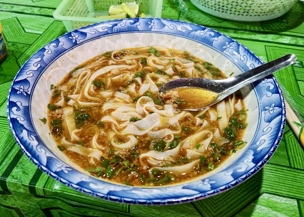 Laos or Vietnam: How to Choose the Best Country for Your Trip 10 A bowl of Khao soi or a local noodle soup is in a blue and white printed bowl sitting on a table with a lime green patterned table cloth at the night market in Luang Namtha, Laos. The soup has a silver soup spoon sititng in it and is filled with wide rice noodles, ground beef, and lots of herbs.