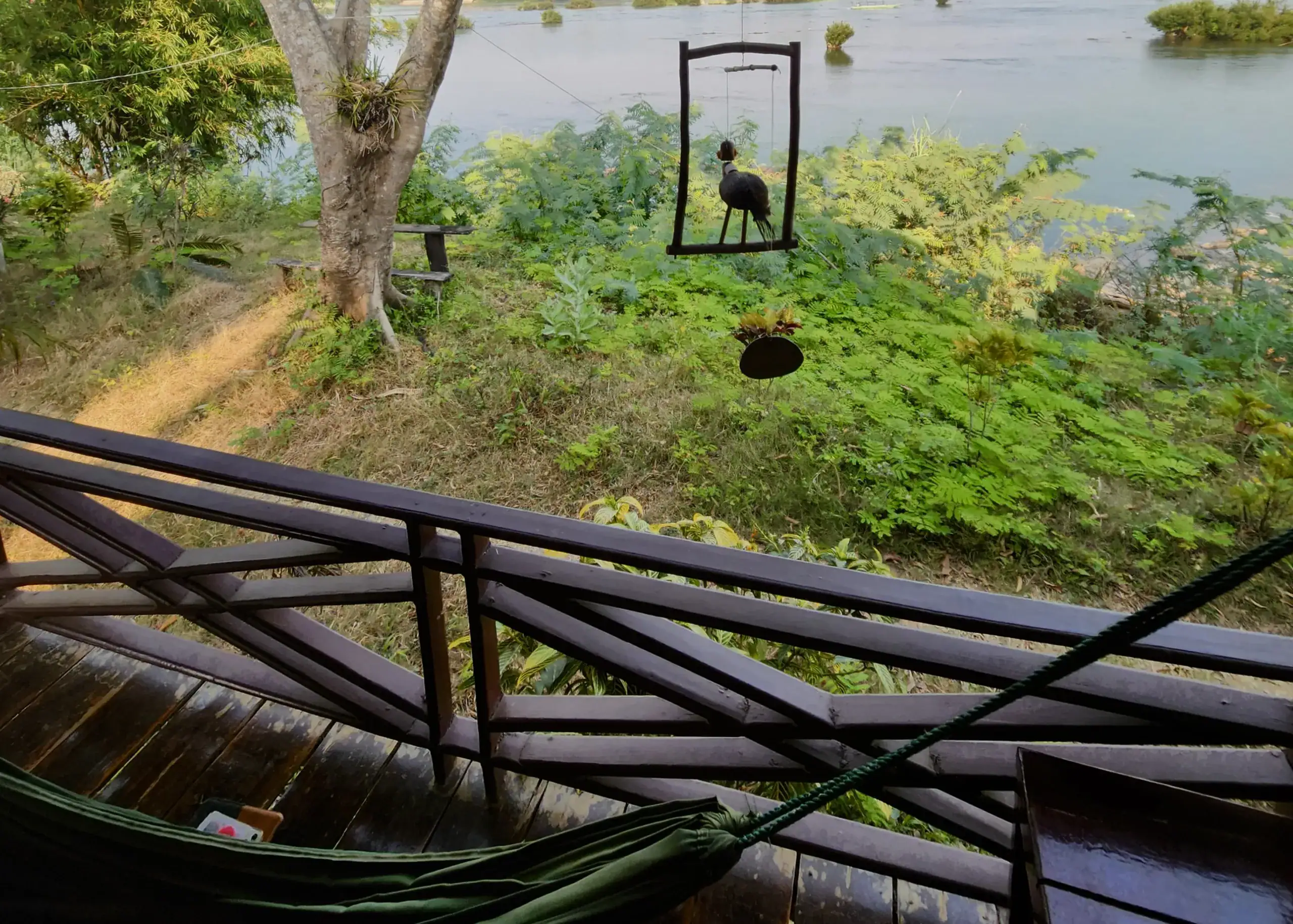 The balcony of a bungalow overlooking the Mekong river in Don Det. There is a hammock and a decoration of a bird hanging from the ceiling of the bungalow
