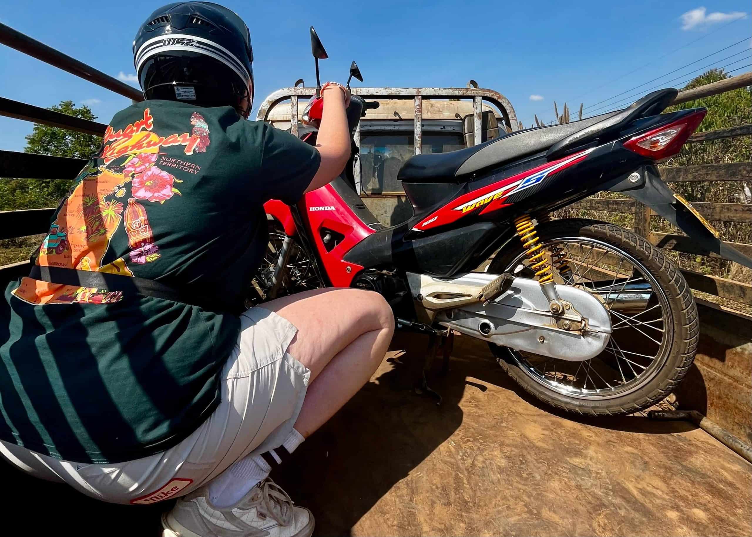Motorbiking in Laos: Routes, Rentals, Costs & Safety Tips 10 A girl in a green t shirt is sitting on the back of a truck holding onto her motorbike that has broken down on the Bolaven loop.