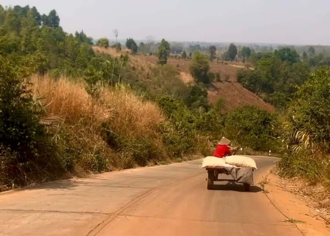 A red dusty road in Laos on the Bolaven Loop. The view is of the road with a man driving a Laotian tractor ahead. THere are trees lining the roads either side of us