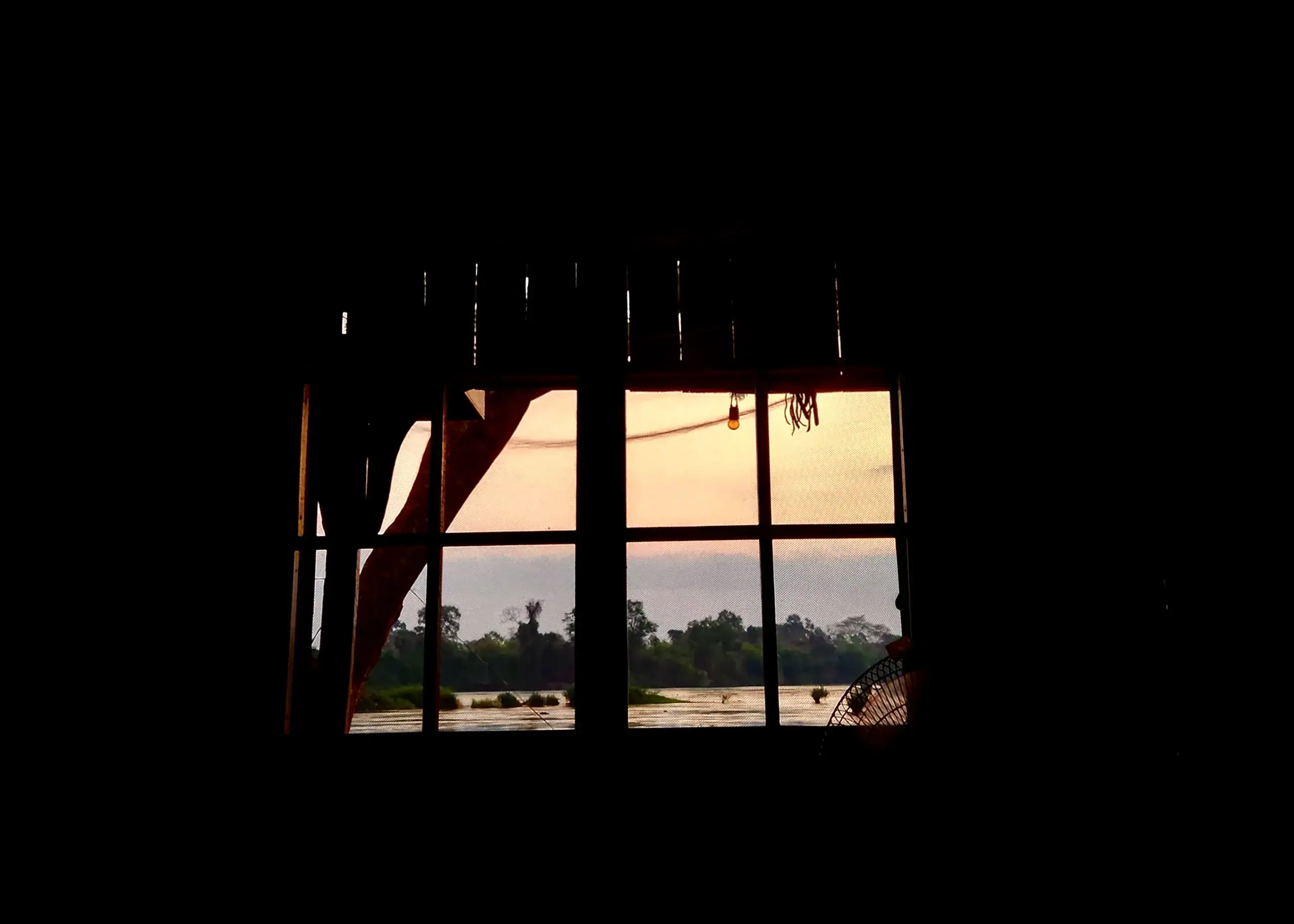 Where to Stay in Don Det & the 4000 Islands, Laos 2 A view of the light pink and orange sunrise from the open window of a bungalow at Mama Leuah guesthouse, a place to stay in Don Det. The scene around the window is still dark apart from light peeking through the wooden slats of the bungalow above the window. You can see the Mekong River and some trees on a small island from the window.
