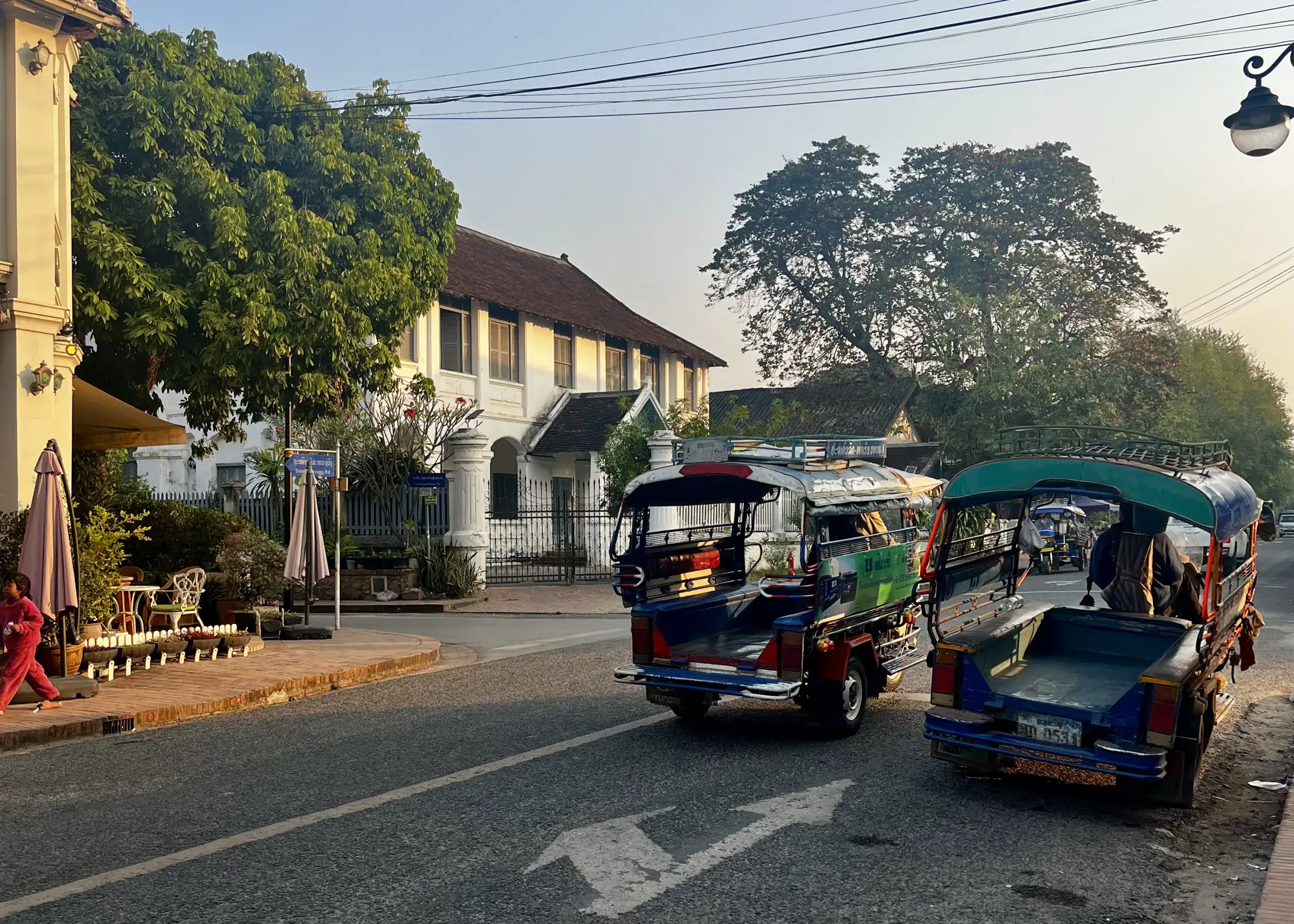Two songthaews parked up on a street in Luang Prabang while the sun is setting. They are motorbikes or pick up trucks converted to a taxi with seating platforms that hold 6-8 people in the back
