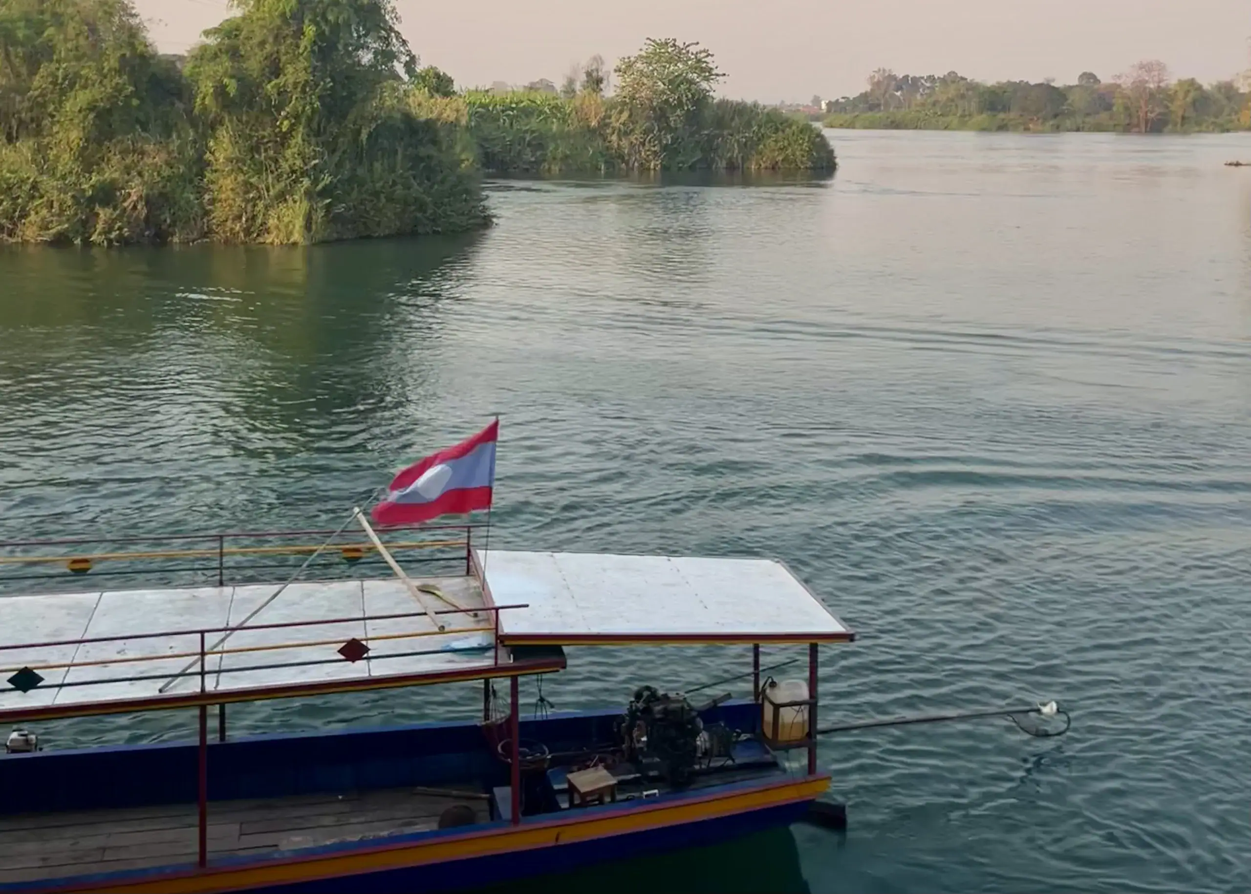 30-Day Laos Itinerary: Unveiling Southeast Asia's Hidden Gem 11 A long boat flying a Laotian flag is floating on the calm, blue Mekong River in Don Det, also called the 4000 Islands, in Laos. The sunset behind the boat is light pink and there is tons of greenery on the little islands dotting the water.