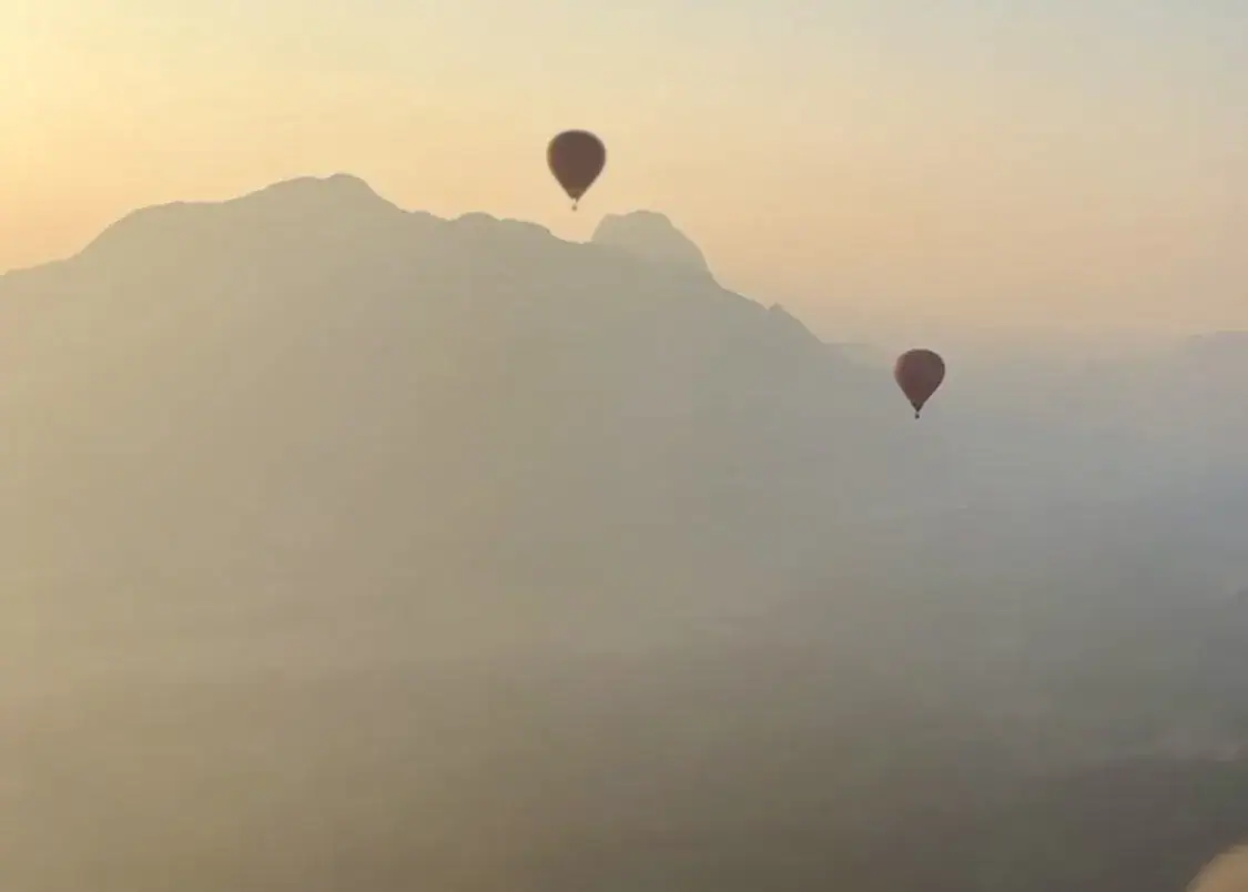 Two hot air balloons floating over the mountains at sunset in Vang Vieng, Laos. The air is hazy and the outline of the mountains is unclear.