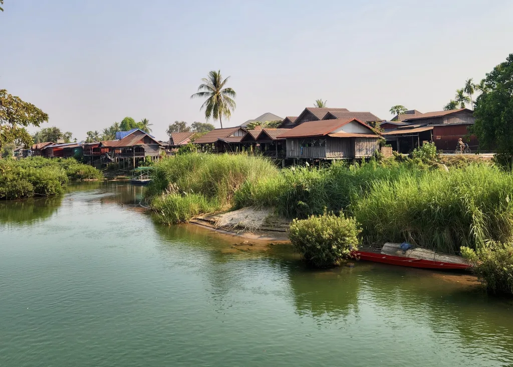 Laos or Vietnam: How to Choose the Best Country for Your Trip 14 A daylight view of the Mekong River in Don Det, Laos. There is a red kayak floating close to the riverbank with a bunch of green scrub on the shore. There are large palm trees and brown wooden bungalows lining the riverbank.