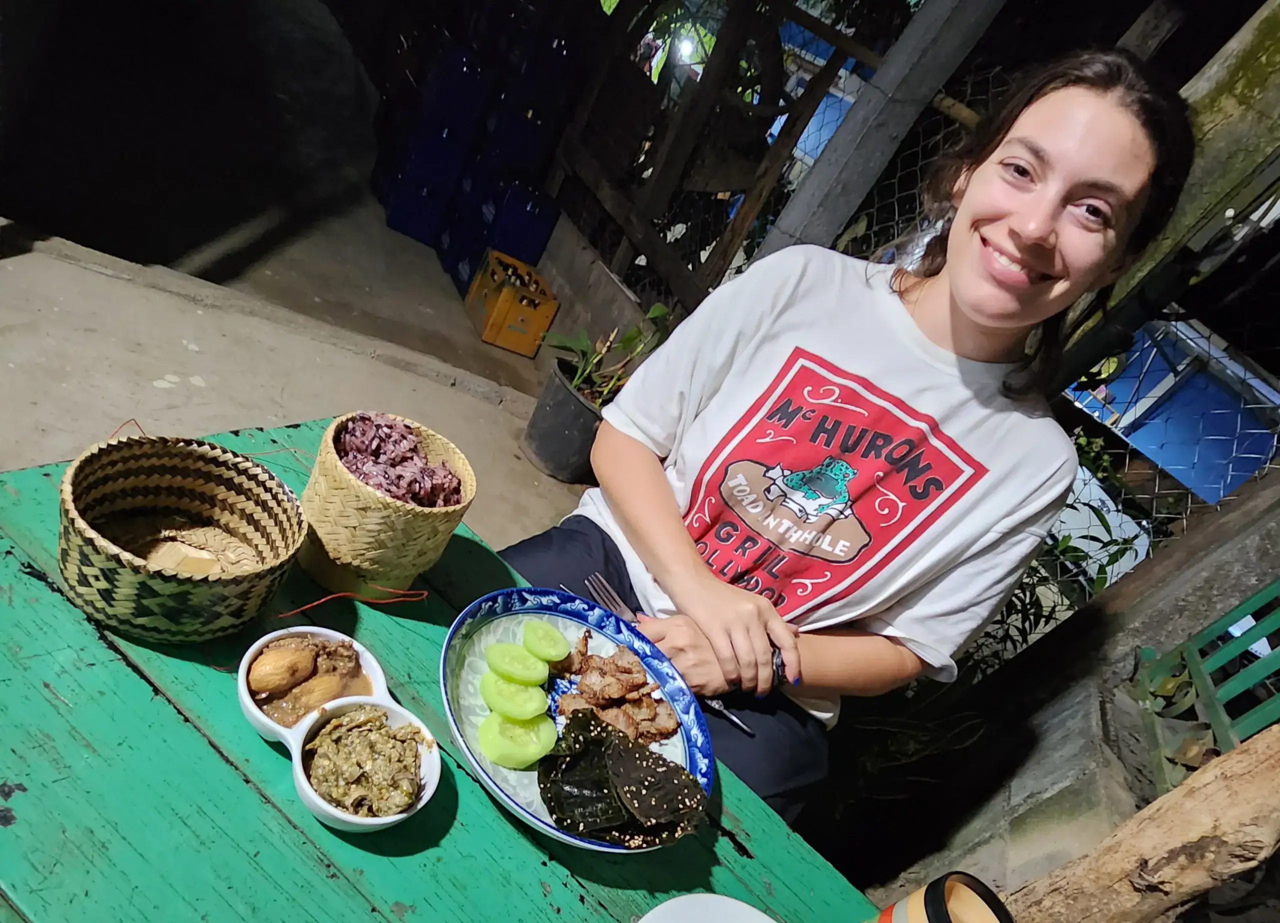 A picture of a girl with brown hair sat at a green table in a restaurant. Her meal is rice in a bomboo container, cucumber, riverweed, eggplant dip and Lao sausage