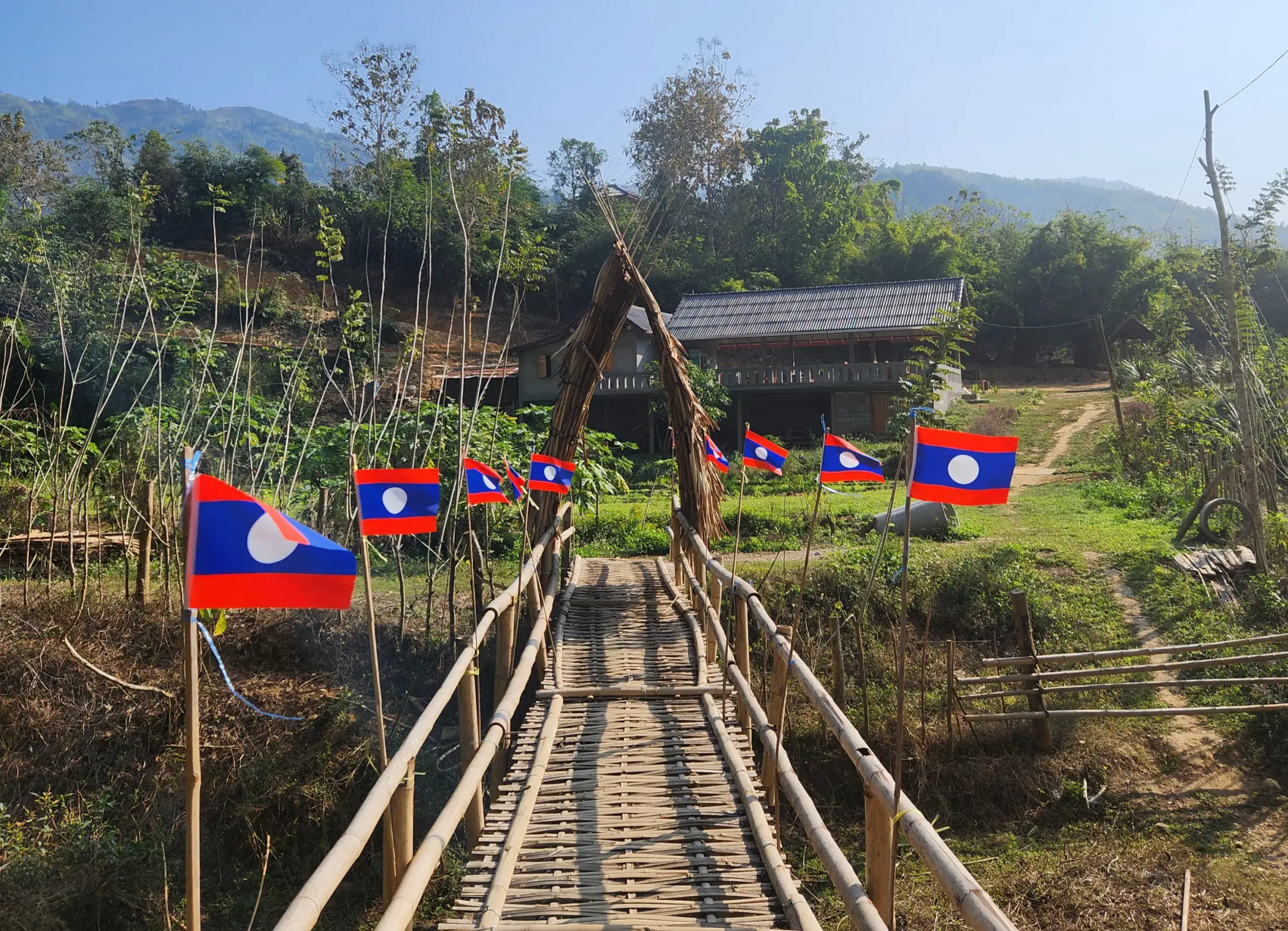 A picture of a bamboo bridge in Nong Khiaw in Northern Laos. The bridge is lined with flags of Laos with lots of greenery in the background