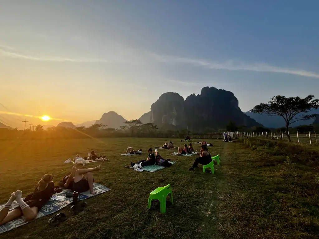 Green chairs lined up in a field with people sat drinking, watching the sunset over the mountains in Laos