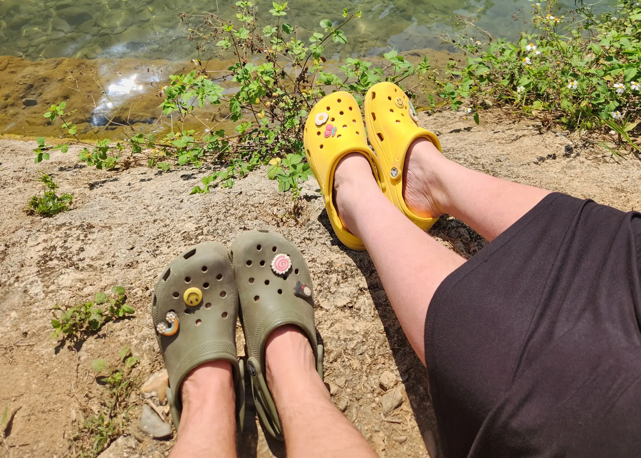 43 Thoughtful and Practical Gift Ideas for Budget Travelers 9 The image shows crocs - the perfect gift for budget travelers. Two people have their feet out on a river bank. One wears green crocs, the other wears yellow crocs