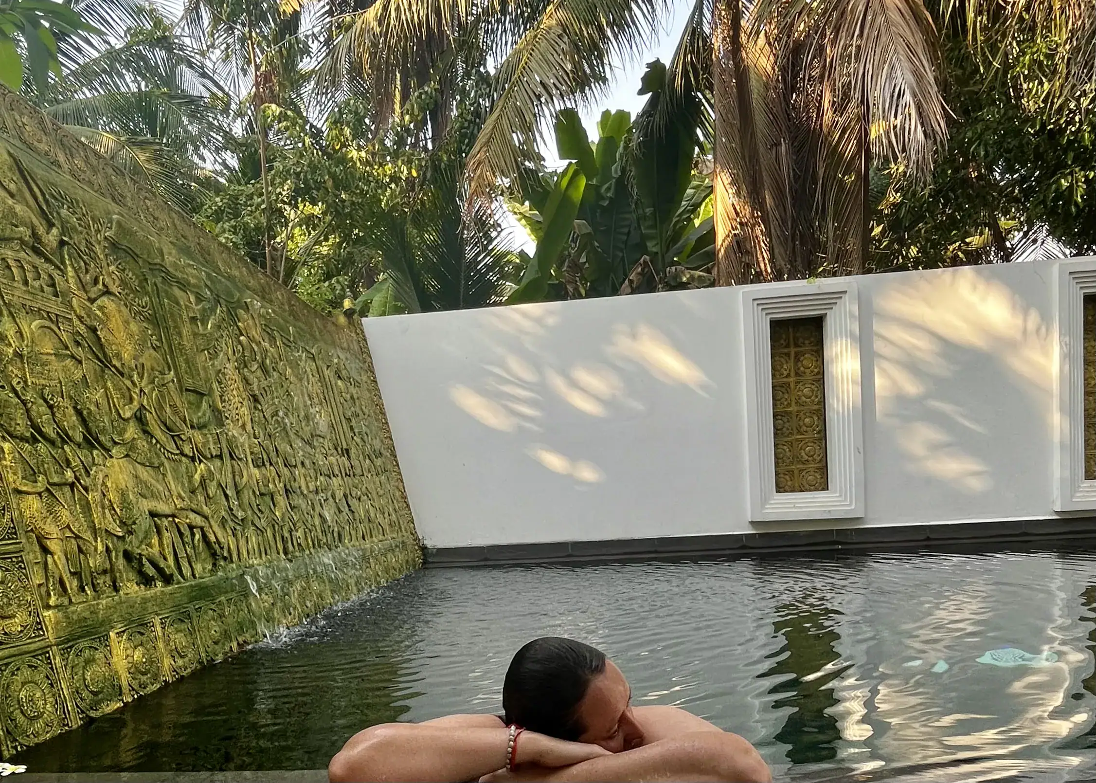 A woman in a swimsuit with wet hair is laying on her crossed arms on the ledge of the pool at The Urban hotel in Siem Reap, Cambodia. One side of the pool is engraved gold figures and the other wall is white with dappled light from the palm trees above the pool shining down.