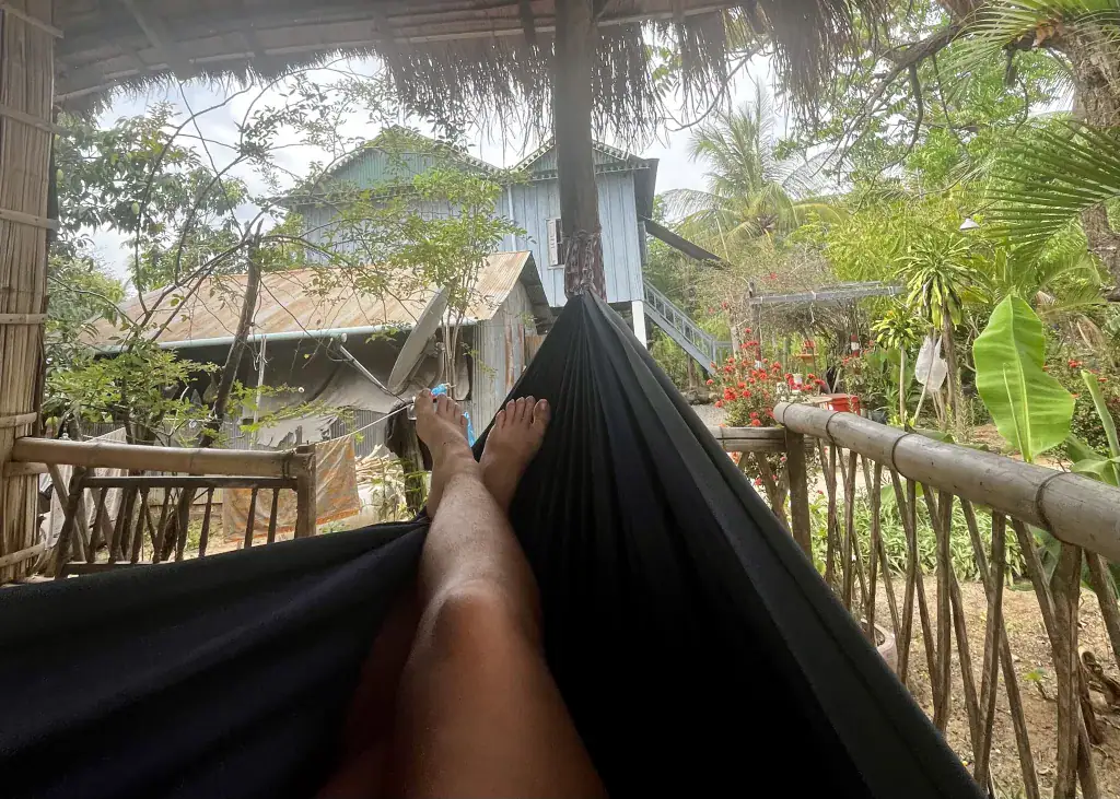 11 Unique Things to Do in Battambang (Even in the Heat!) 6 A person sat in a black hammock on a balcony in Battambang. The view is the bamboo home and the greenery in the garden. The girl is sat cross legged in the hammock