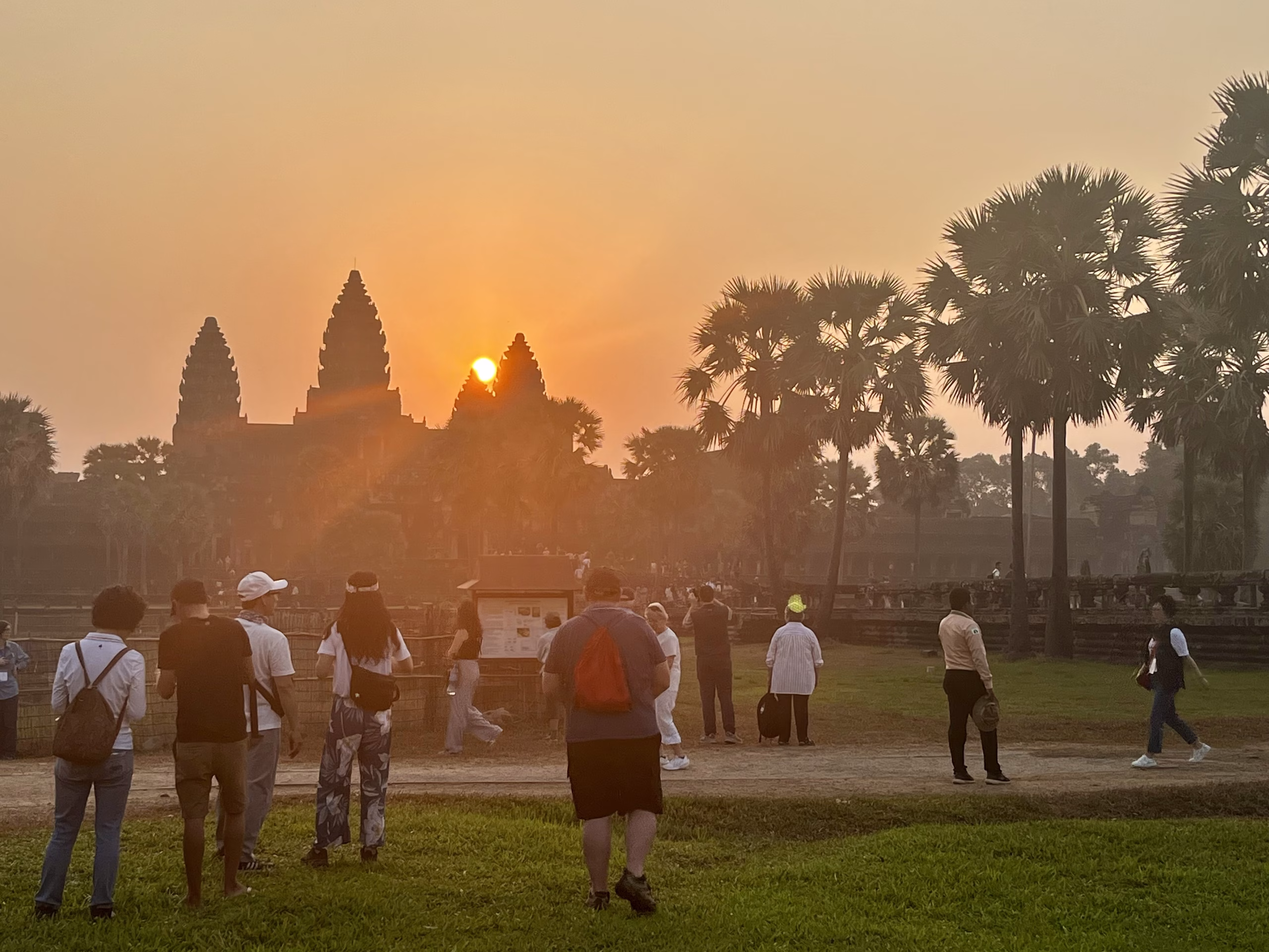 Best Angkor Wat Tour for Sunrise - Our Top Picks 3 The sun rising above the temple on an Angkor Wat sunrise tour. There are people standing on the grass taking photos.