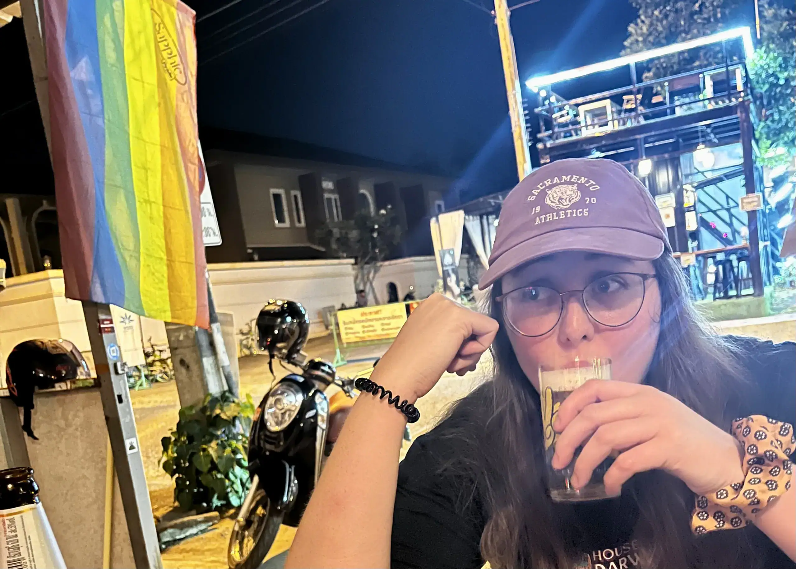 Chiang Mai or Chiang Rai: Which Is Better? 2 A girl in a red hat, wearing glasses is sipping a beer outside a bar called Sapphic Riot in Chiang Mai. There is a LGBTQ+ flag hanging in front of the bar