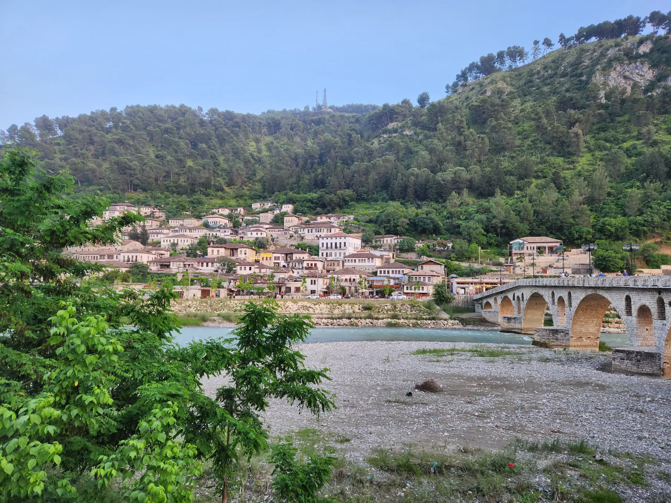 2 Weeks in Albania Without a Car: Mountains, Beaches & Hidden Gems 9 The picture is taken in Berat, as part of an Albania Itinerary. There are white houses stacked on top of each other in the background, then a river running through the middle, with pebbles on the other river bank