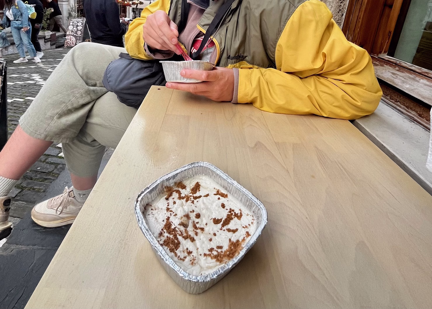 There are two people sat at a small table on a street. The girl taking the picture has a pudding like dessert in front of her in a square foil tray, with cinnamon on top. The girl across from her is wearing a yellow coat and is eating the dessert with a spoon