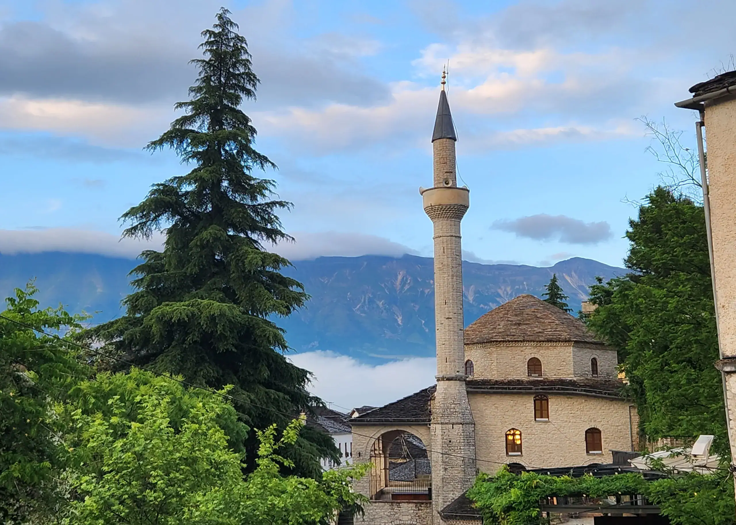 Albania 9 A typical view in Gjirokaster, Albania. There are mountains lined with clouds in the background. In the foreground is a pine tree on the left, then a slightly smaller mosque to the right of the tree. This is a must-stop destination on an Albania itinerary 7 days.