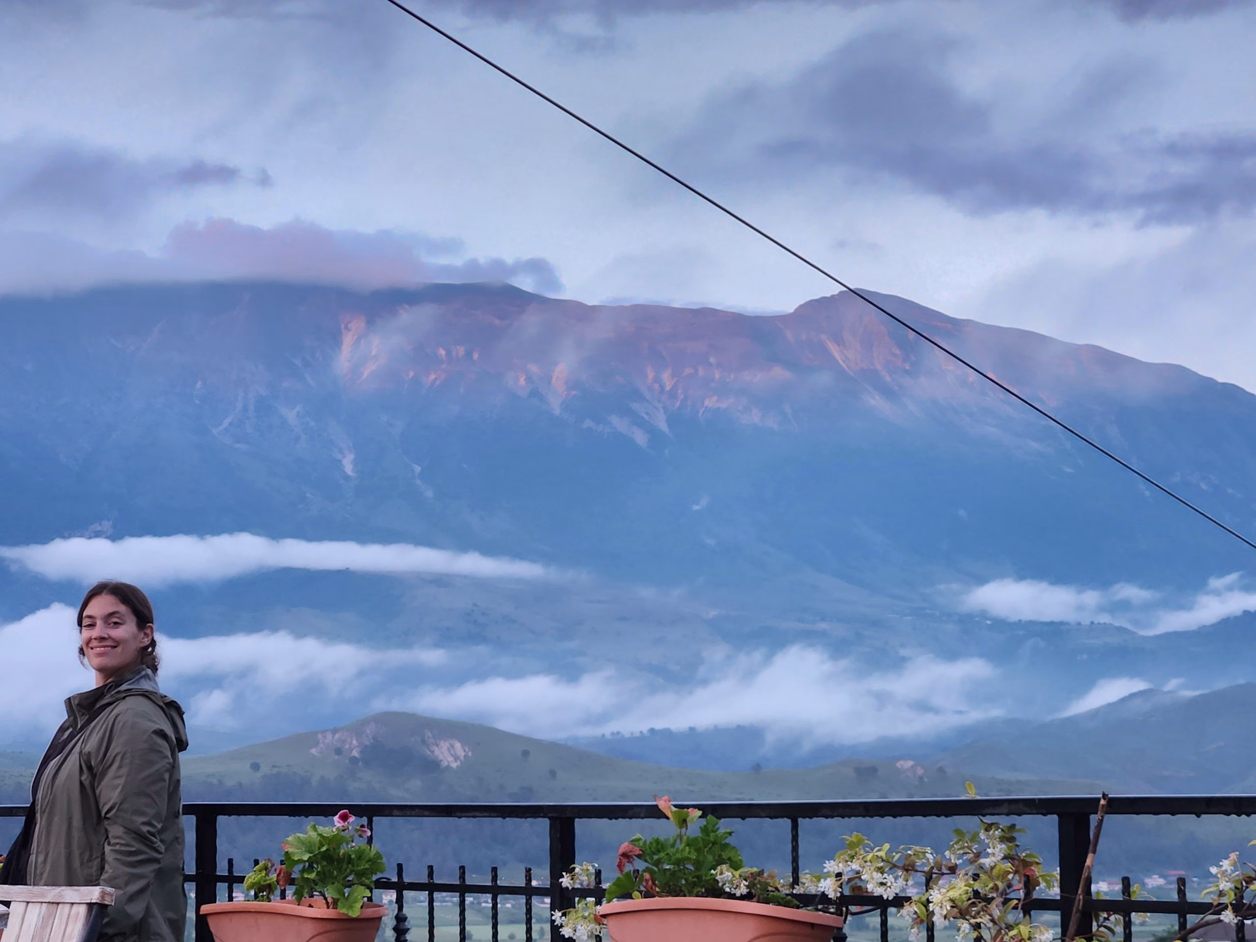 A picture of a girl in a green coat stood on the balcony of Friends Hostel in Gjirokaster. The black fence is lined with terra-cotta plant pots and colourful flowers. In the background is the montains that surround Gjirokaster, with low cloud sitting across them.