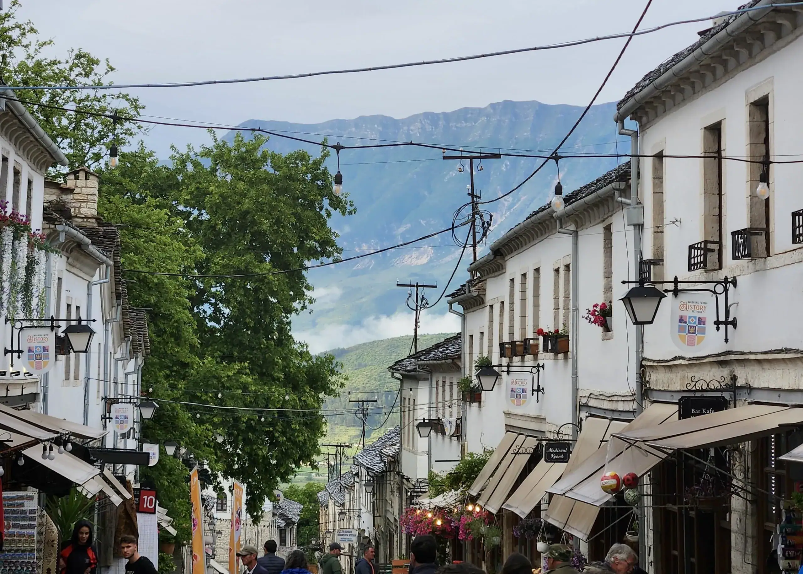 2 Weeks in Albania Without a Car: Mountains, Beaches & Hidden Gems 11 Old white buildings line the cobbled streets of Gjirokaster, Albania. There are mountains in the background and telephone wires connecting all the buidlings. It is foggy because a storm has just cleared.