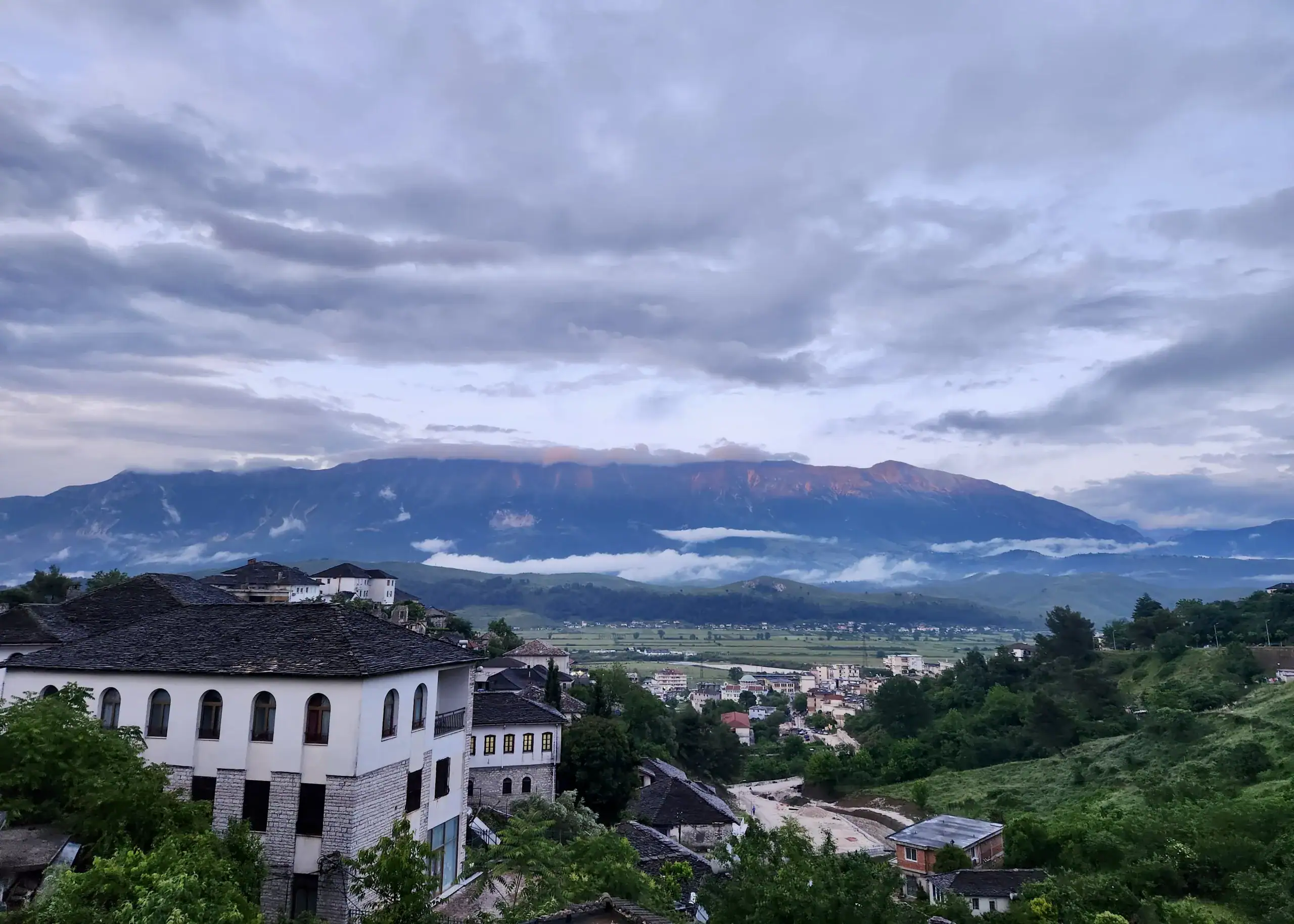 Albania 1 The city of Gjirokaster, Albania is covered in a light fog at sunset. There are a lot of green trees on the hills below the mountains and white buildings in the distance. Be sure to visit if you have 2 weeks in Albania.