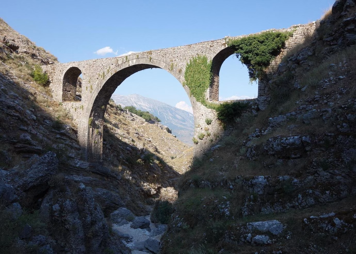 The picture shows Ali Pasha bridge near Gjirokaster. The tall bridge is made of stone and has a wdie tall archway, built in traditional Ottoman style. It is built over a valley that meets below the bridge