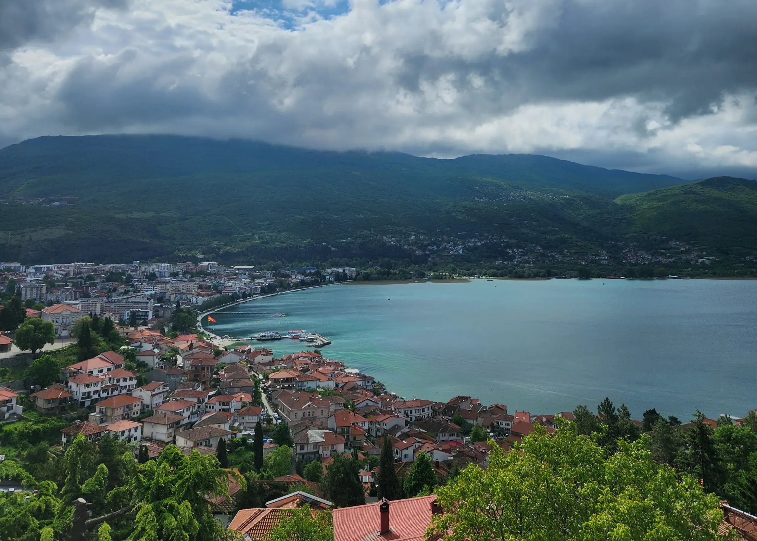 One Week South Albania Itinerary: Beaches, Castles & Culture 13 Views over Lake Ohrid. The blue water is surrounded by mountains, trees and meditteranean houses with orange roof