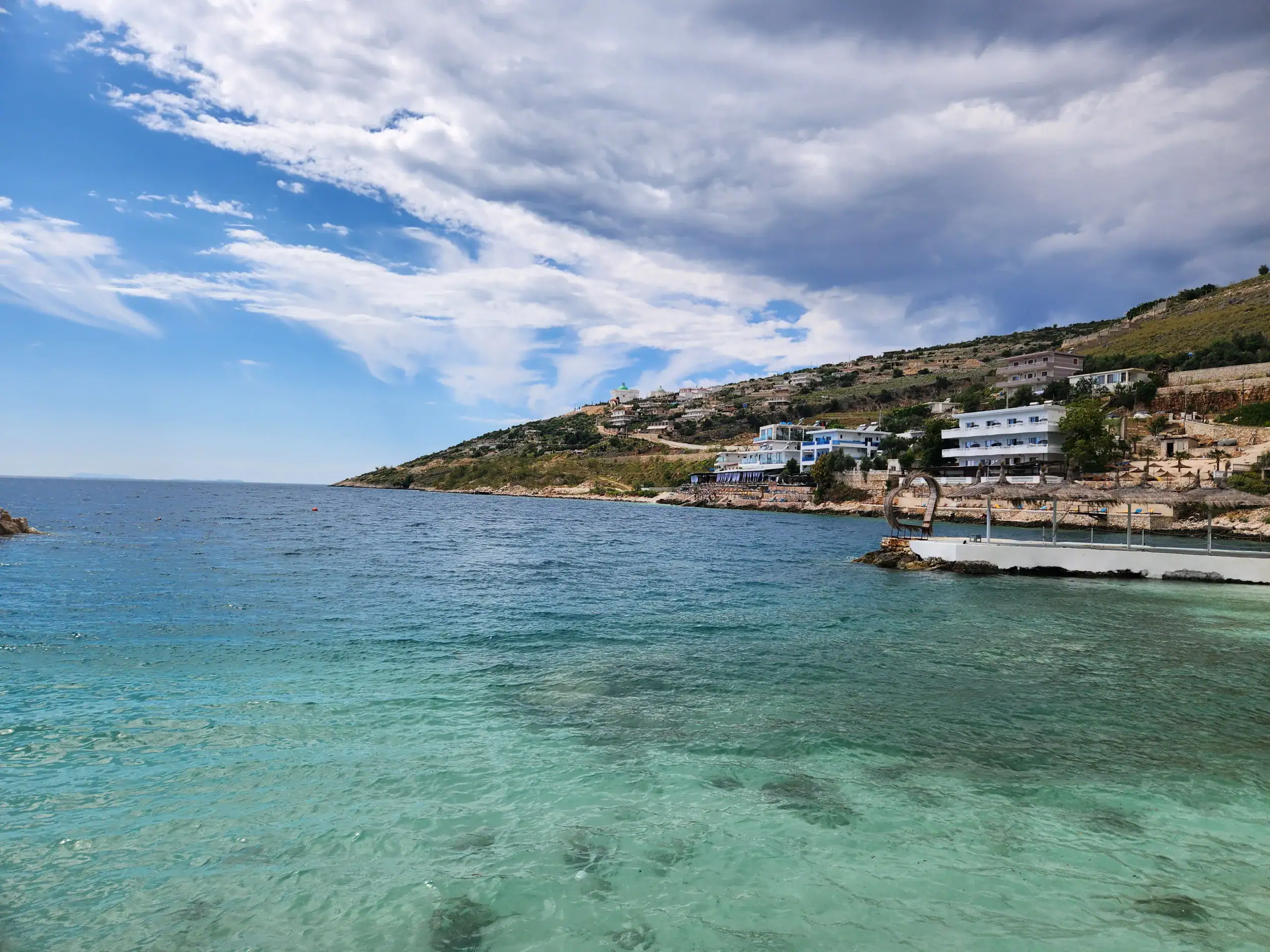 One Week South Albania Itinerary: Beaches, Castles & Culture 6 Clear blue waters fill most of this image. There is a hill with hotels on in the background. The crystal clear waters are in Sarande, a stop during 7 days in Albania