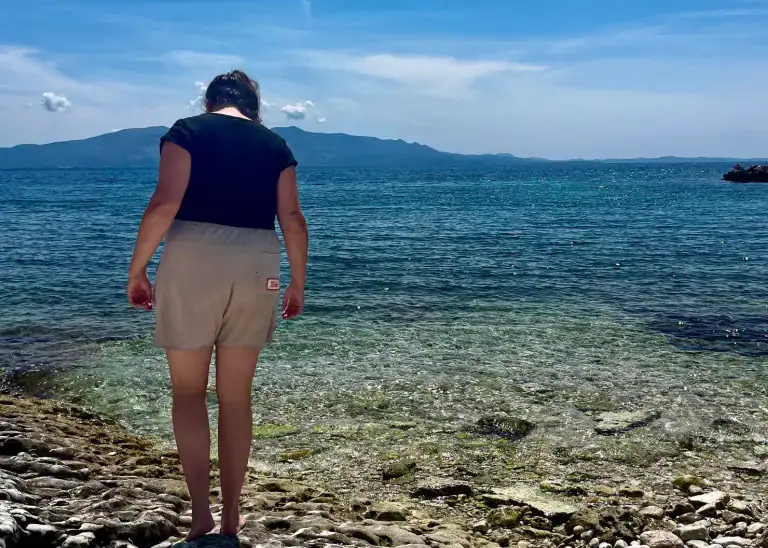 A girl in white shorts and a black tank top is walking on rocks towards a shoreline. The rocks are a light grey and the turqoise sea is crystal clear