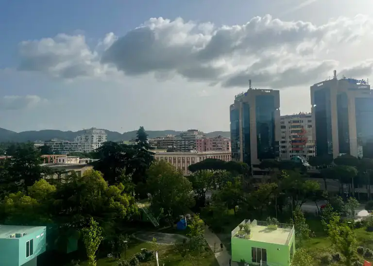 The image shows the city views from the Pyramid of Tirana. You can see green tree tops, two sky scrapers to the right, and lots of houses set in front of mountains in the distance