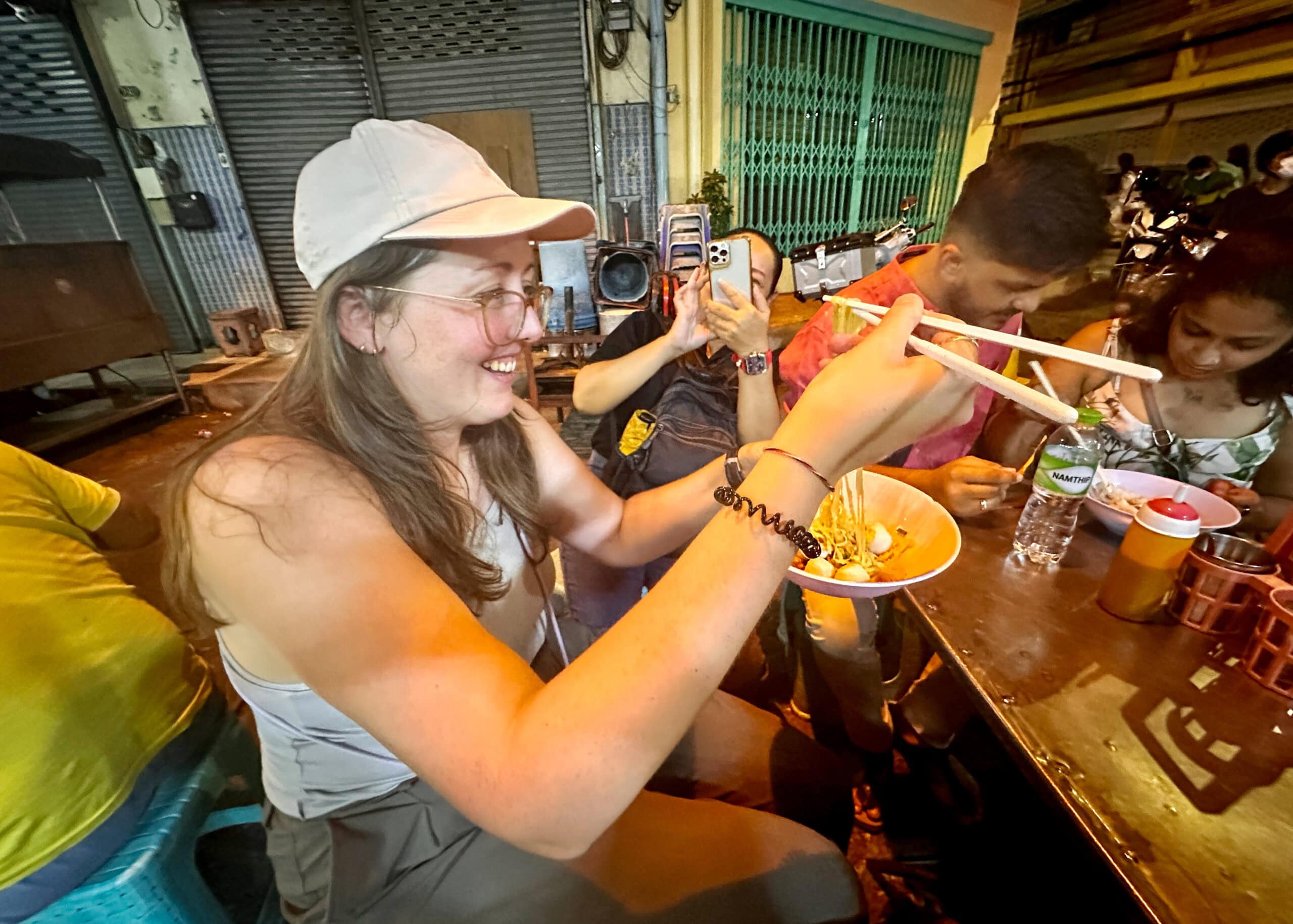 BEST Food in Chinatown Bangkok: What is Actually Worth It 3 Jess is sitting in the alley of a famous Bangkok restaurant. She's sitting on a stool at a metal table, smiling while pulling noodles up from her soup.