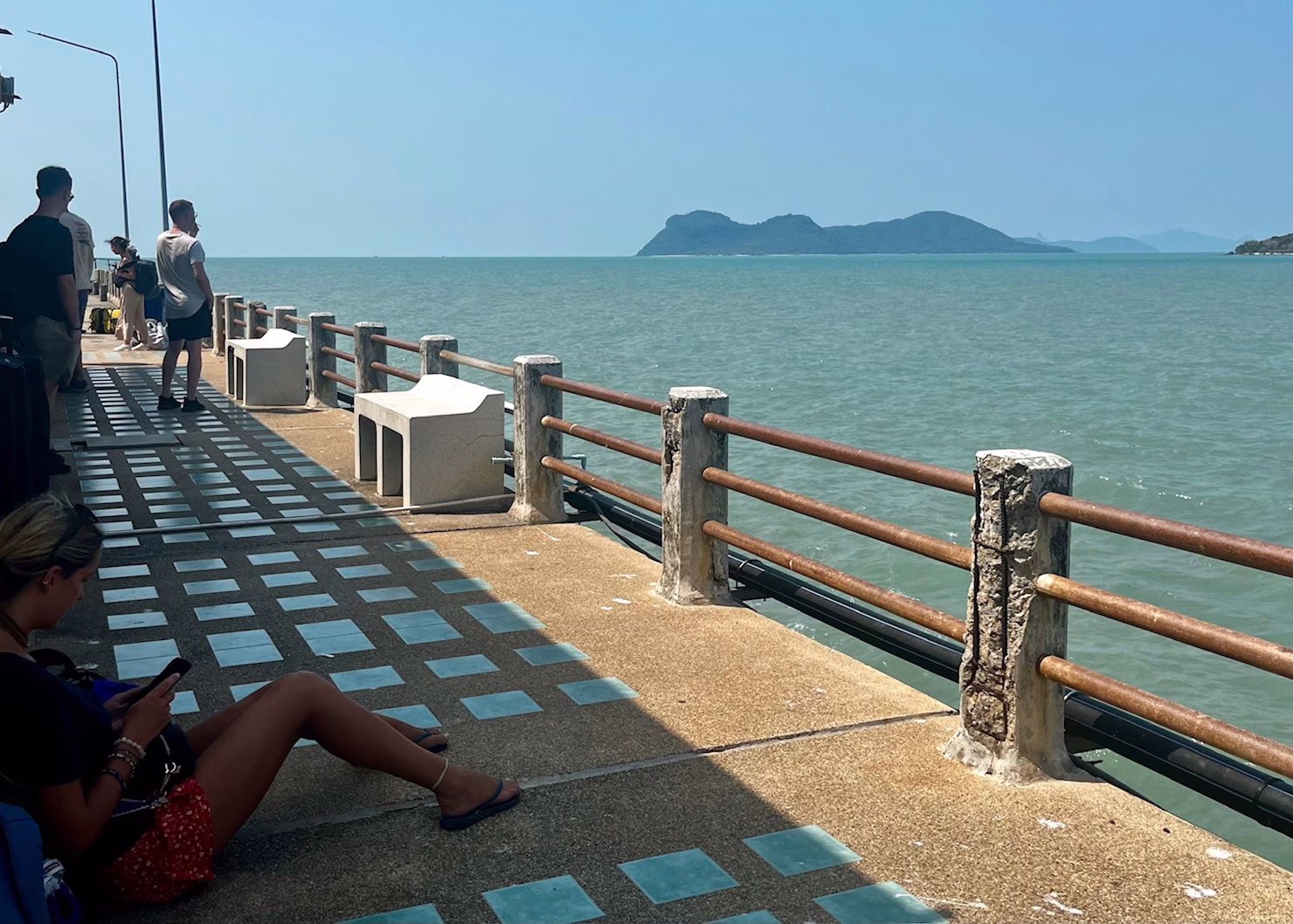 Ferries to Koh Lanta: Routes, Tips & Real Advice 3 A woman is sitting in the shade of a building on a pier that is for ferries heading to Koh Tao. The water under the pier is bright blue and there are distant islands in the background.