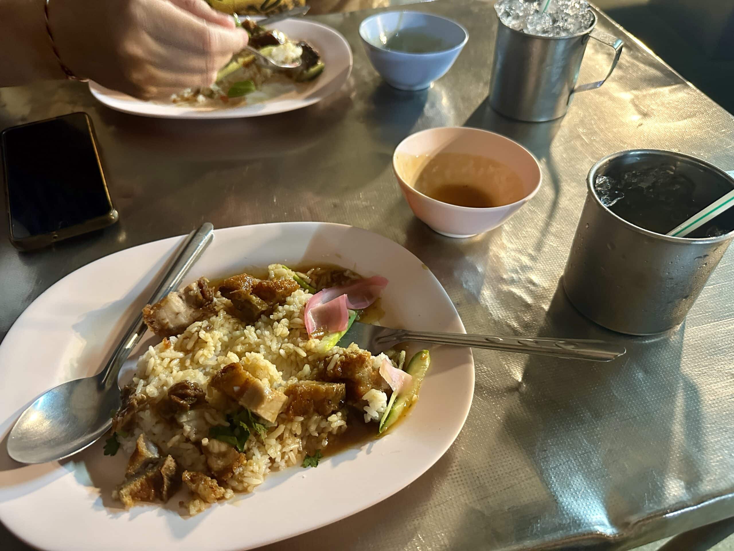 BEST Food in Chinatown Bangkok: What is Actually Worth It 5 A dish of crispy pork and rice with iced tea sits on a classic Thai pink plate on a metal table on the street in Bangkok.