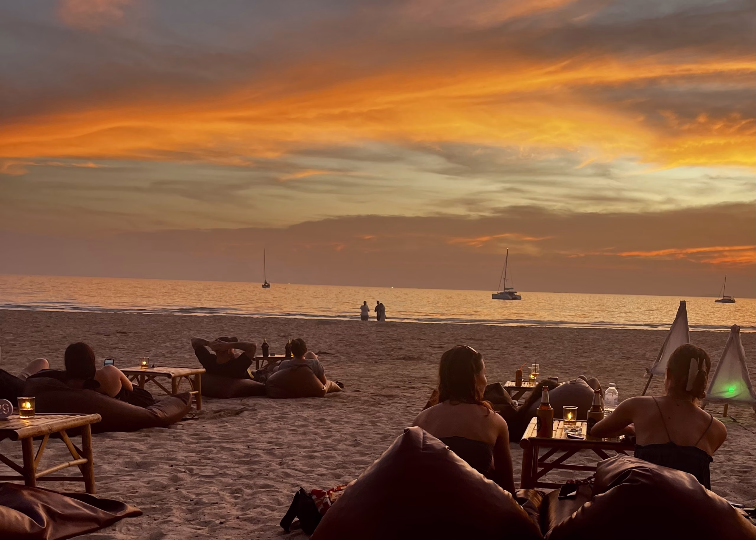 A orange and blue sunset is shown over Long Beach in Koh Lanta. There are visitors sitting on bean bags on the sand with drinks watching the sunset at one of the many restaurants on the beach.