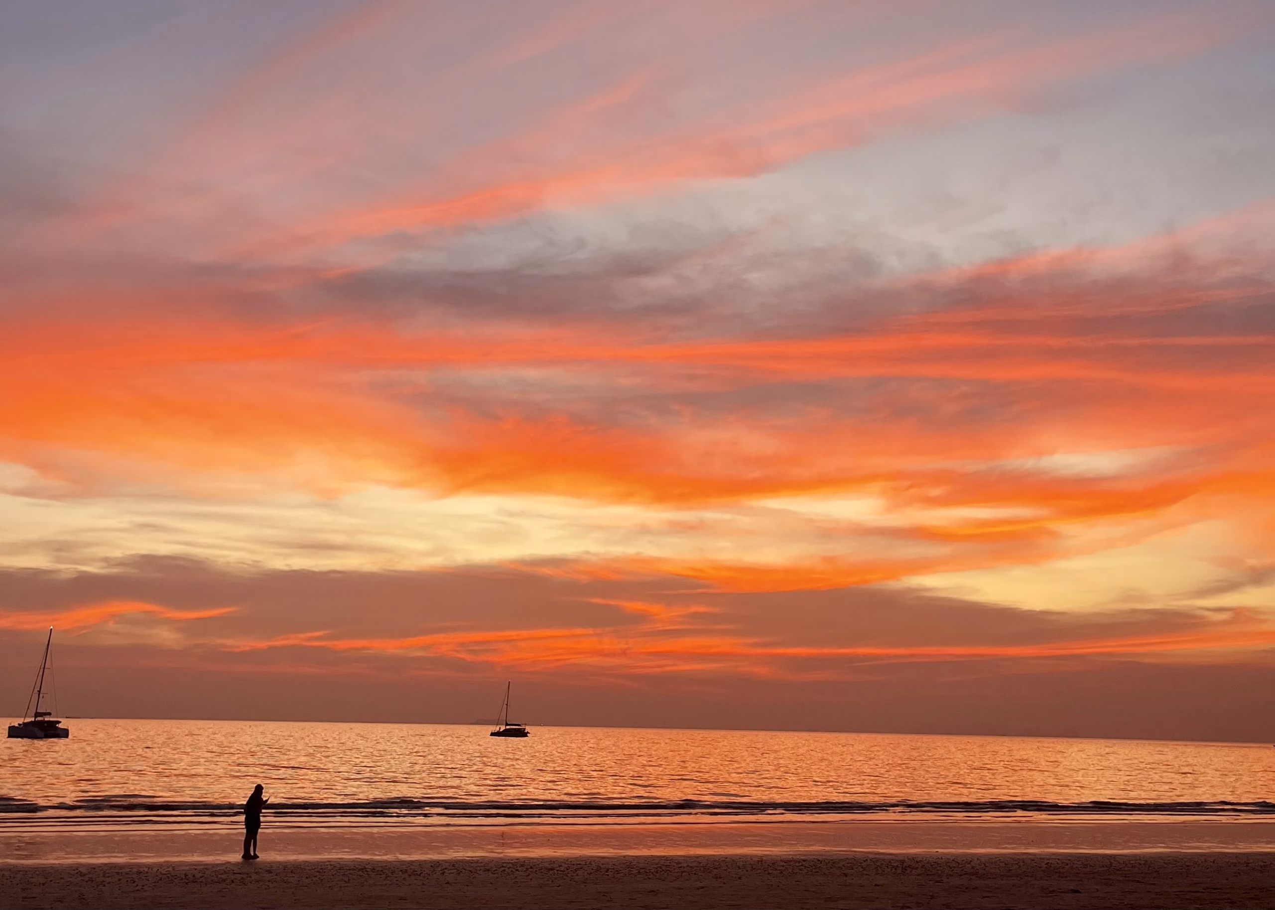 A bright orange and pink sunset over Long Beach in Koh Lanta, Thailand. There is a dark outline of a sailboat on the water and a person walking on the beach in the distance.