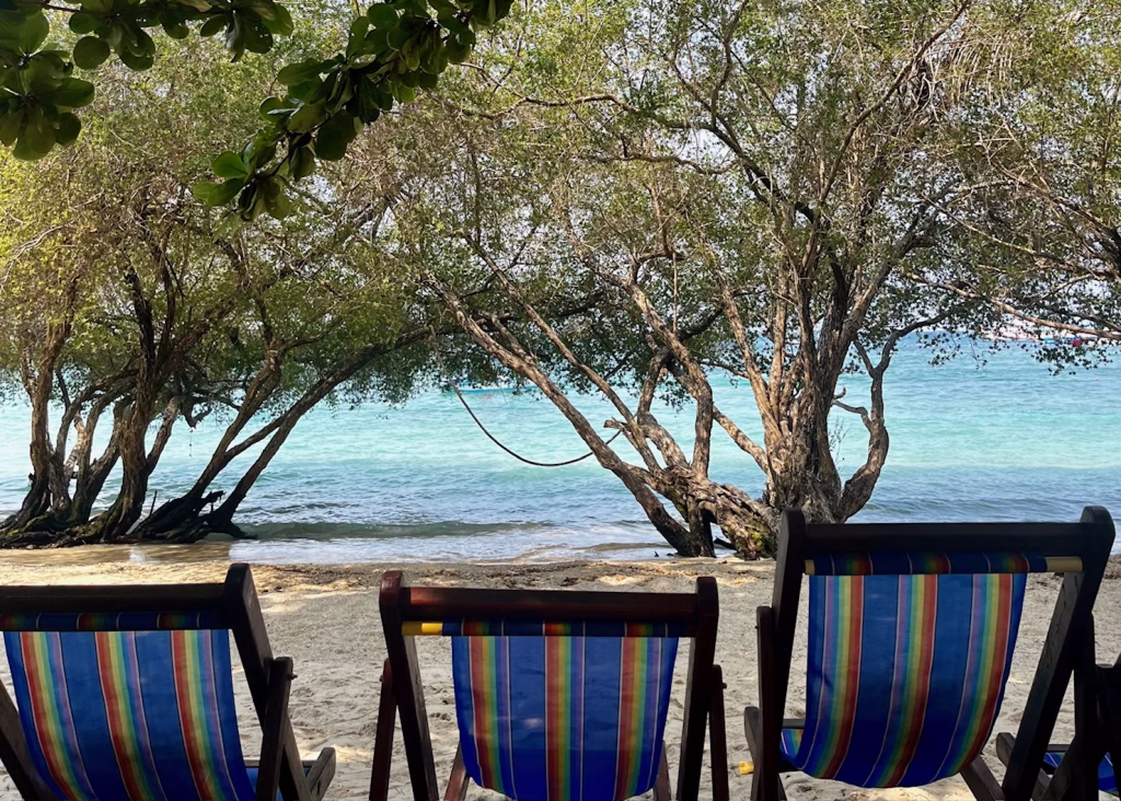 Where to Go in Thailand for the First Time: A Budget Traveler’s Guide 7 Three striped rainbow beach chairs are sitting on tan sand at Freedom Beach in Koh Tao. The blue water and clear sky is partially shielded by big trees, which give the beach shade during the day. This is an easy beach to reach if you're visiting Koh Tao without a scooter.