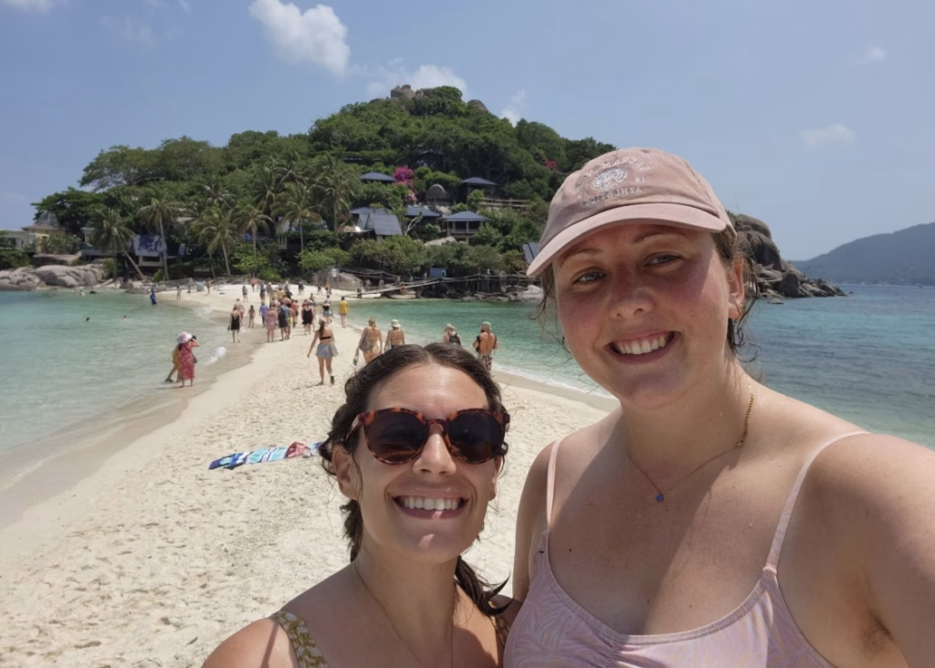 Best Snorkeling in Koh Tao: Local Tips, Top Tours & Hidden Gems 2 A lesbian couple is posing for a picture together at a stop on a snorkeling tour from Koh Tao. There is a mountain with green shrubs, white sand, and blue water behind them.