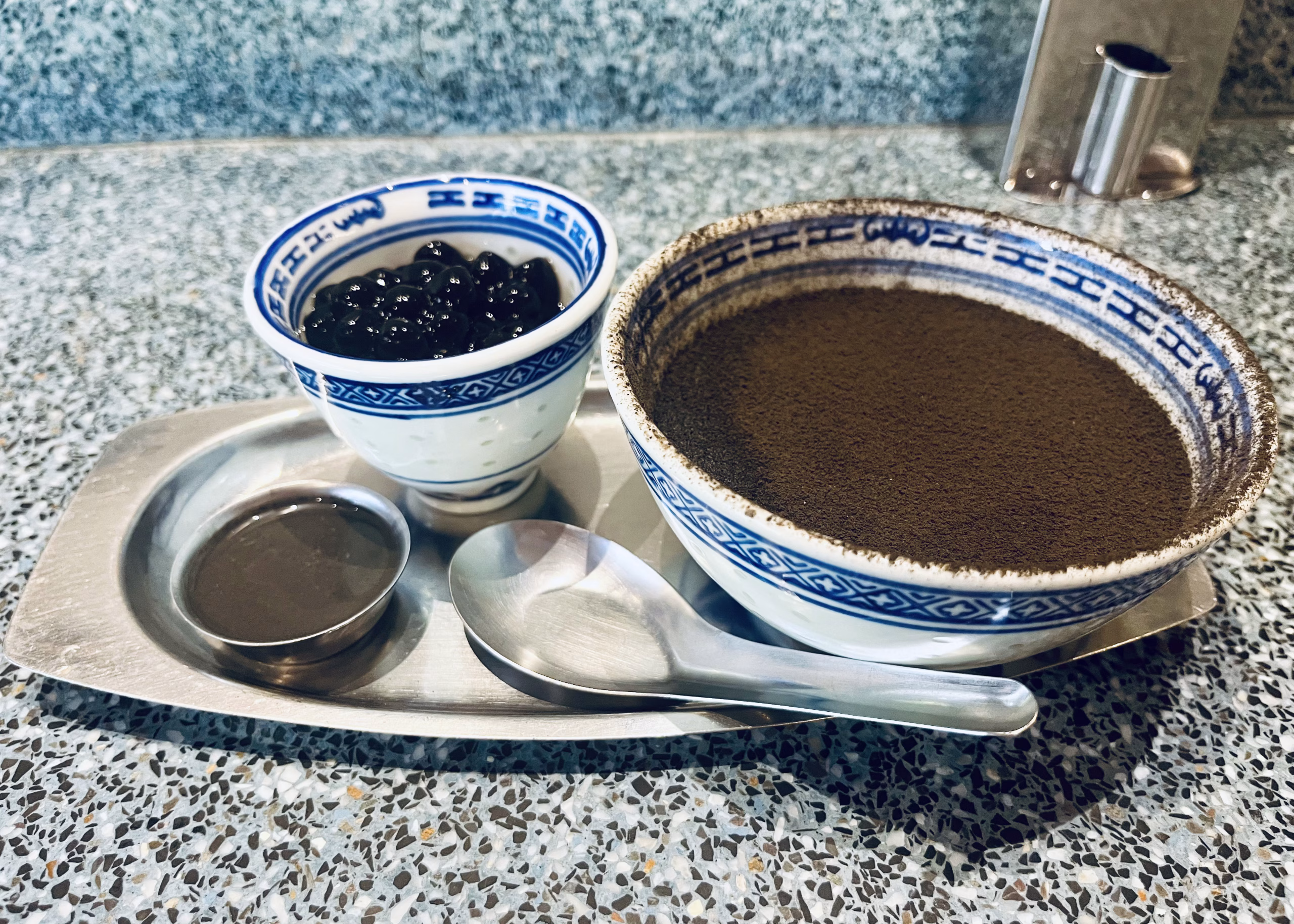 BEST Food in Chinatown Bangkok: What is Actually Worth It 10 Blue and white glass bowls are sitting on a silver serving tray with a silver spoon. In the bowl is a black sesame soy pudding with boba balls and condensed milk to put over top of the pudding.