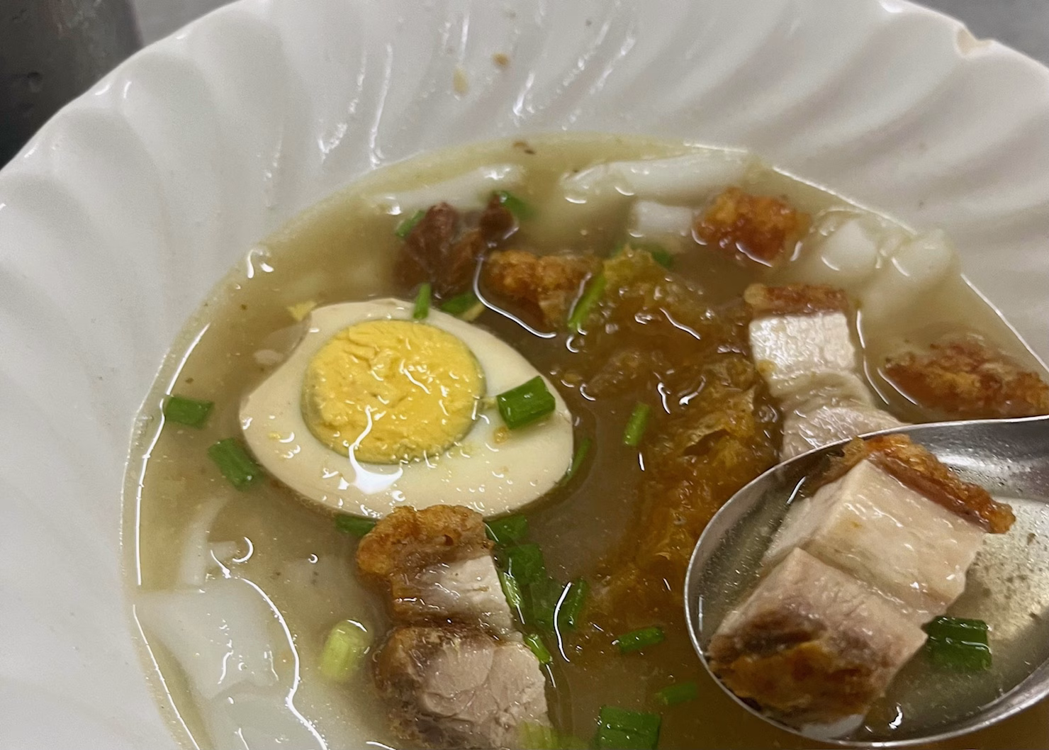 BEST Food in Chinatown Bangkok: What is Actually Worth It 4 A white bowl of noodle soup with pork belly bits, scallions, and a boiled egg are sitting on a table in a hole in the wall restaurant in Chinatown Bangkok.