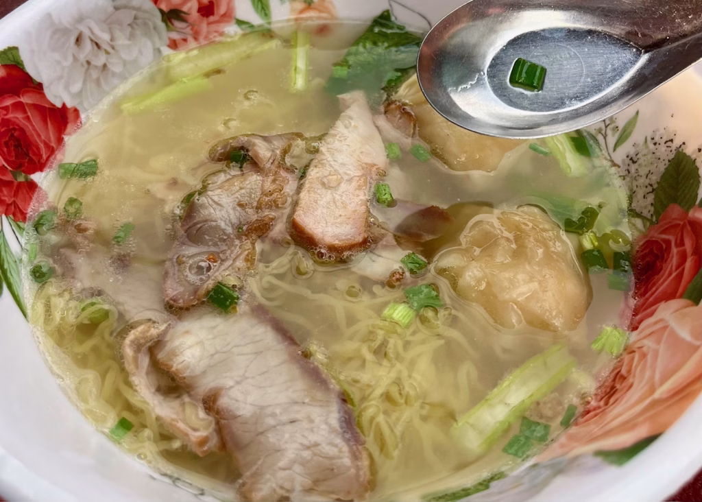 A bowl of thin noodles with slices pork and dumplings, with some green bok choy are in a white and pink flowered bowl. There is a silver metal spoon sitting on the side of the bowl at a fishball stand in Chinatown Bangkok.