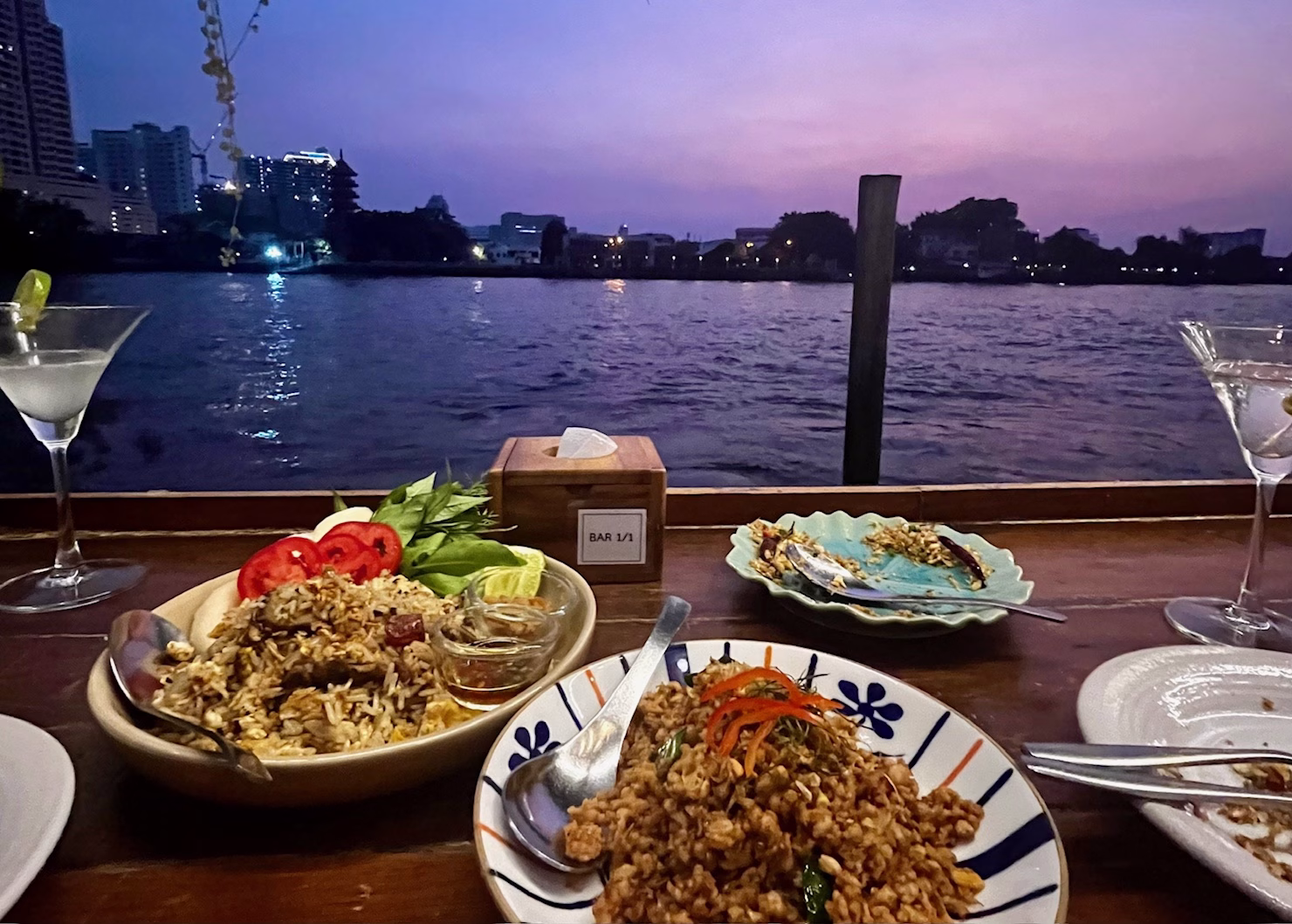 BEST Food in Chinatown Bangkok: What is Actually Worth It 6 Two plates of thai food are sititng on a wooden bar that overlooks the river in Chinatown, Bangkok. The sunset is a deep purple and pink hue and there are a few boats lit up on the river.