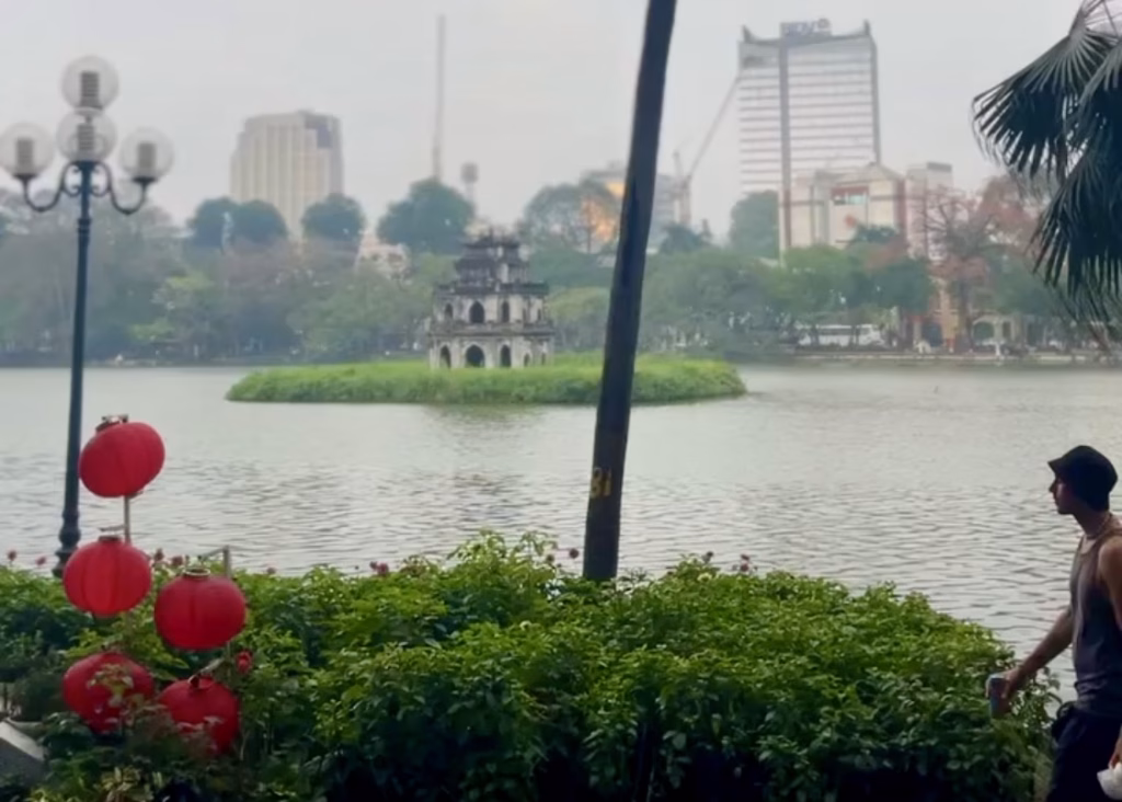 Where to Stay in Hanoi: Best Neighborhoods + Our Favorite Hotels 3 A misty evening along Hoàn Kiếm lake in Hanoi features Ngoc Son Temple in the middle of the lake. There is a tall palm tree in the center of the photo as well as red flowers along the shore contrasting with the gloom of the city and weather.