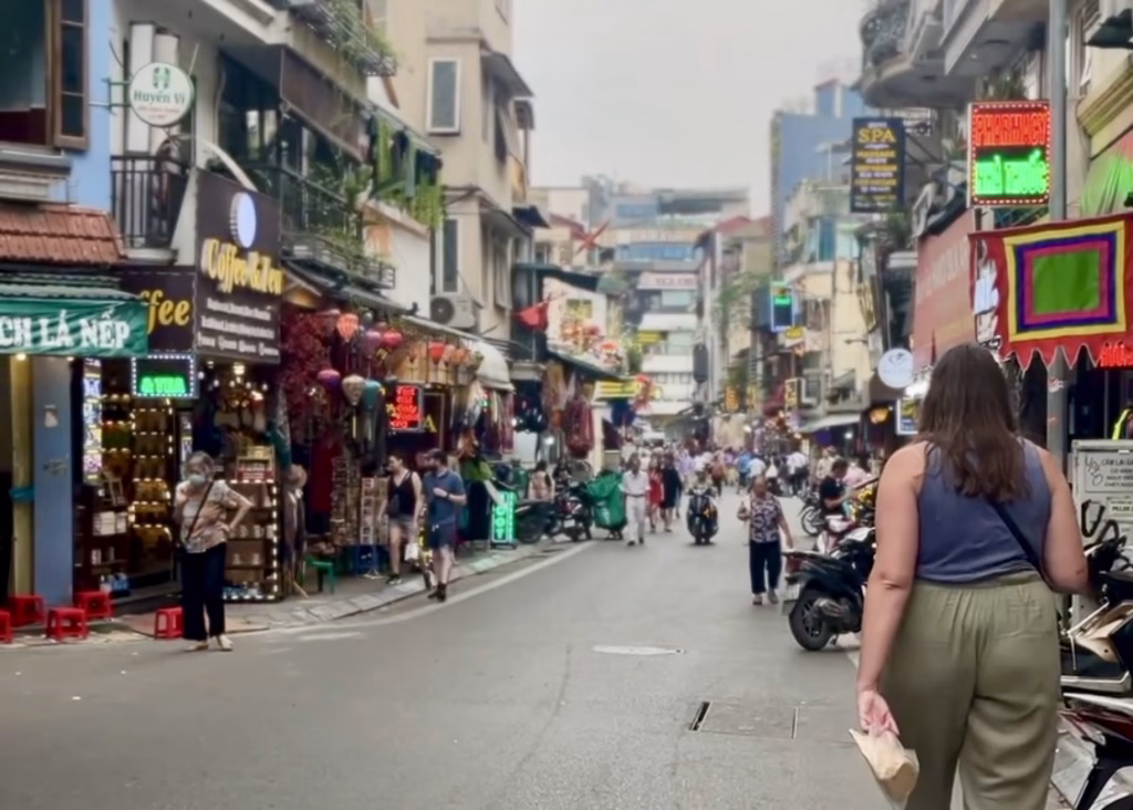 Laos or Vietnam: How to Choose the Best Country for Your Trip 7 A woman in green pants and a blue vest top is walking on the side of a busy street in Hanoi. She is carrying a bag of donuts and there are buildings built up and neon lights on either side of her.