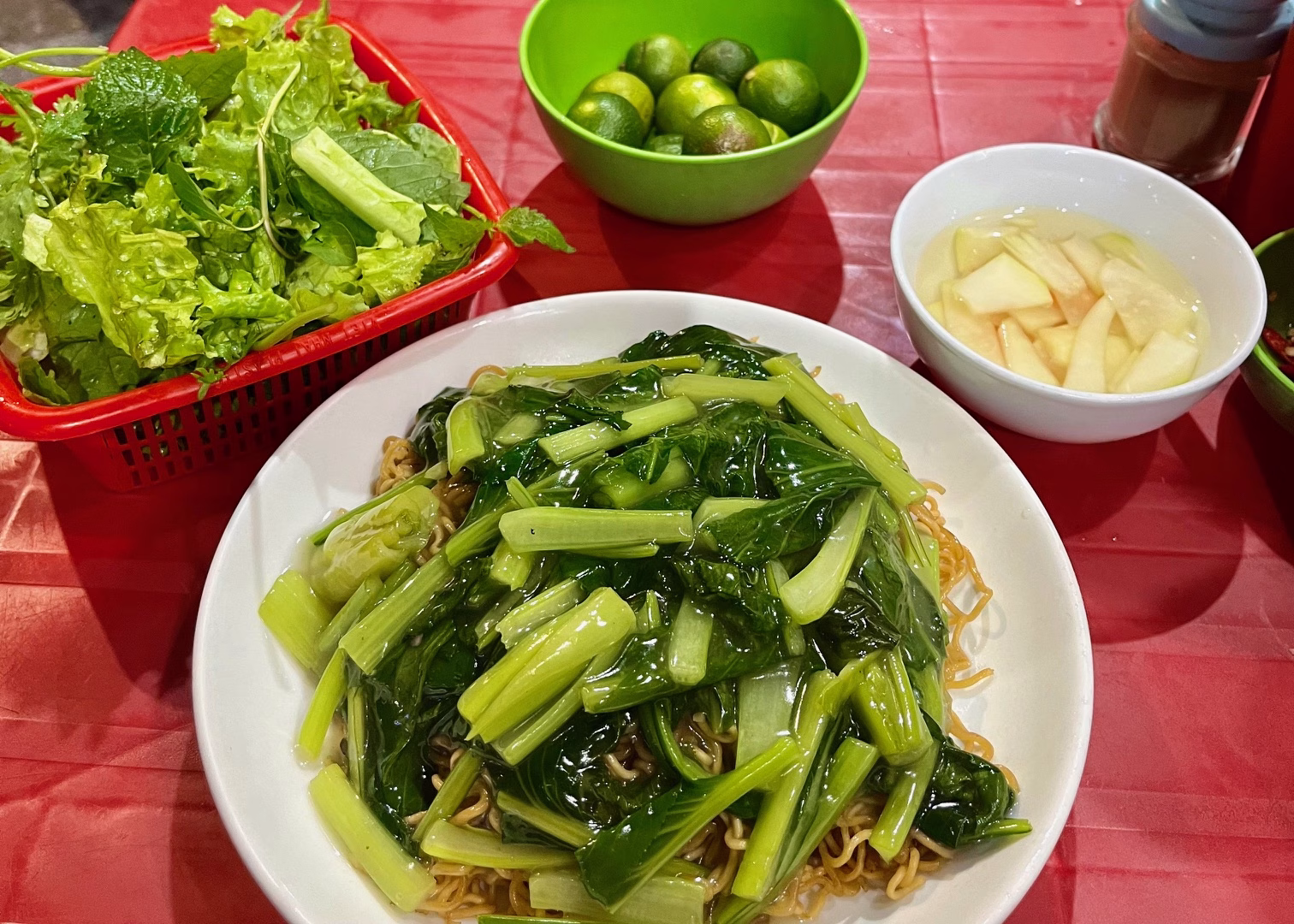 Food in Hanoi: The Best Old Quarter Restaurants 11 A plate of vegetarian food is sitting on a red table. The plate has noodles sitting under a pile of fried morning glory. There is a red basket of herbs, a green bowl of limes, and a bowl of ginger sitting next to the main dish.