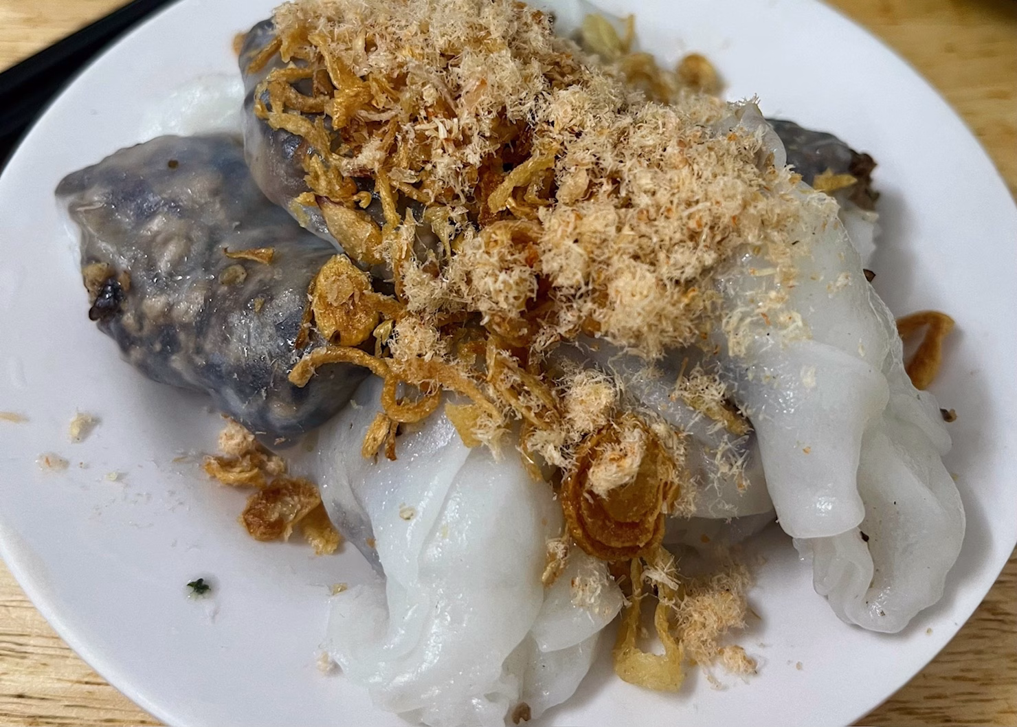 Food in Hanoi: The Best Old Quarter Restaurants 5 Two Vietnamese rice rolls are sitting on top of each other on a white plate that is sitting on a wooden table. The rice rolls are filled with pork and mushrooms which show through the wrapper. There are brown crispy fried scallions and pork floss on top of the rolls. This is some of the best food in Hanoi and is often included in food tours here.